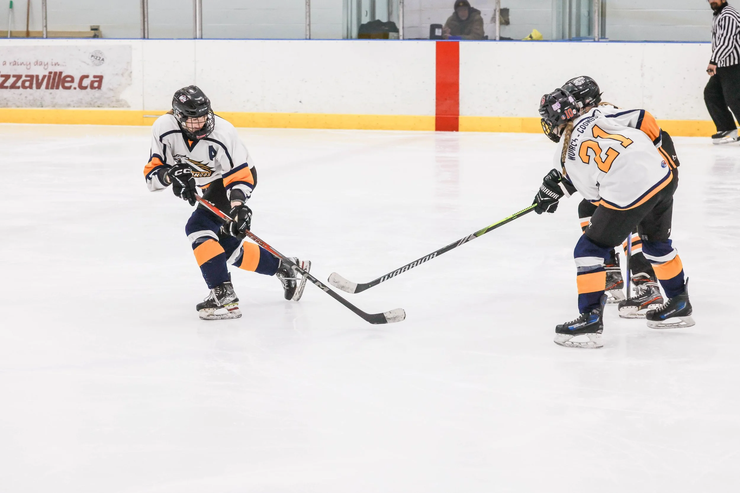 Two young hockey players in white jerseys with orange and navy accents compete for the puck on the ice rink, with a referee in the background.