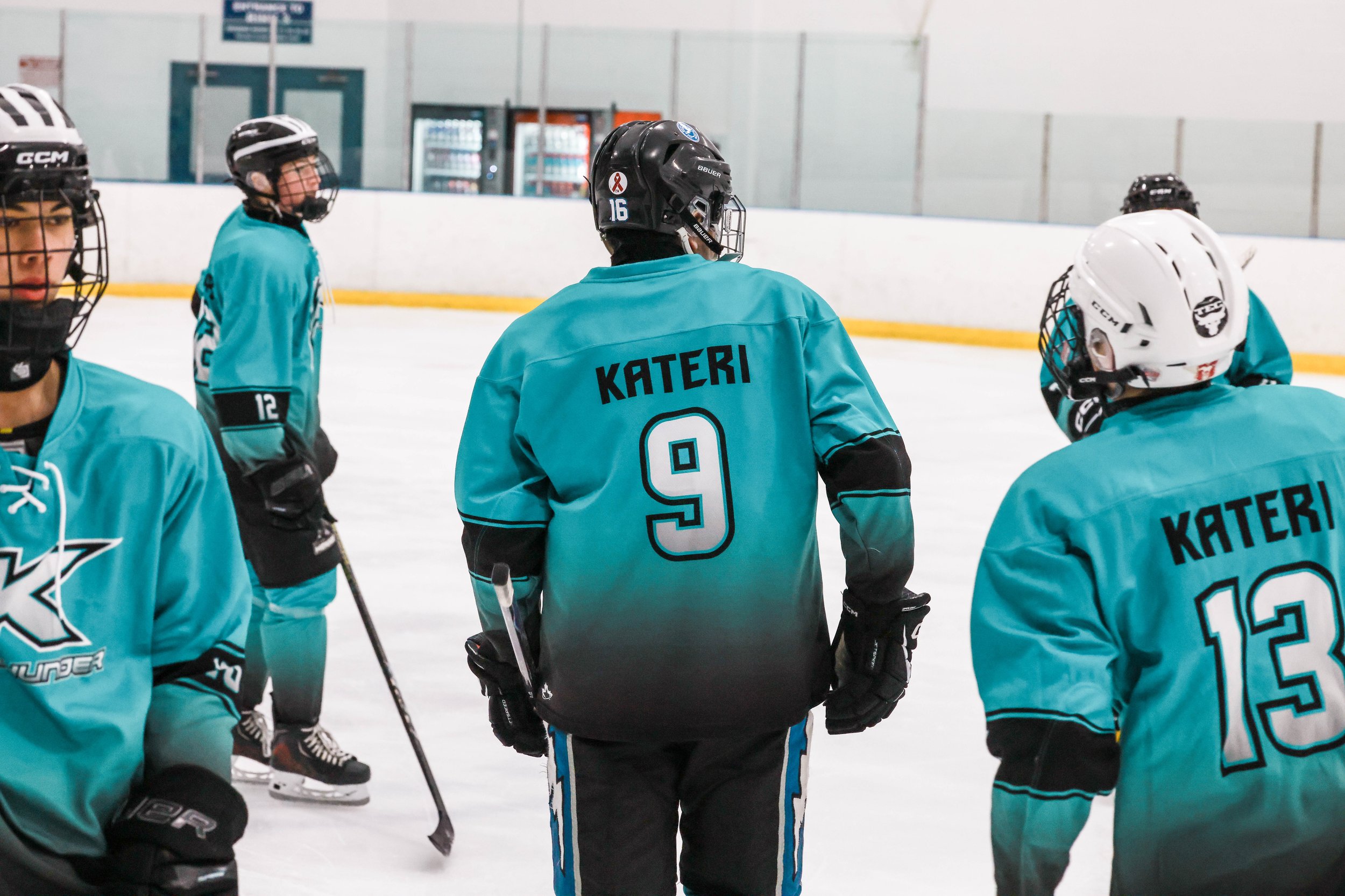 Group of young hockey players on an ice rink wearing teal jerseys with the name 'KATERI' and numbers 9 and 13 visible.