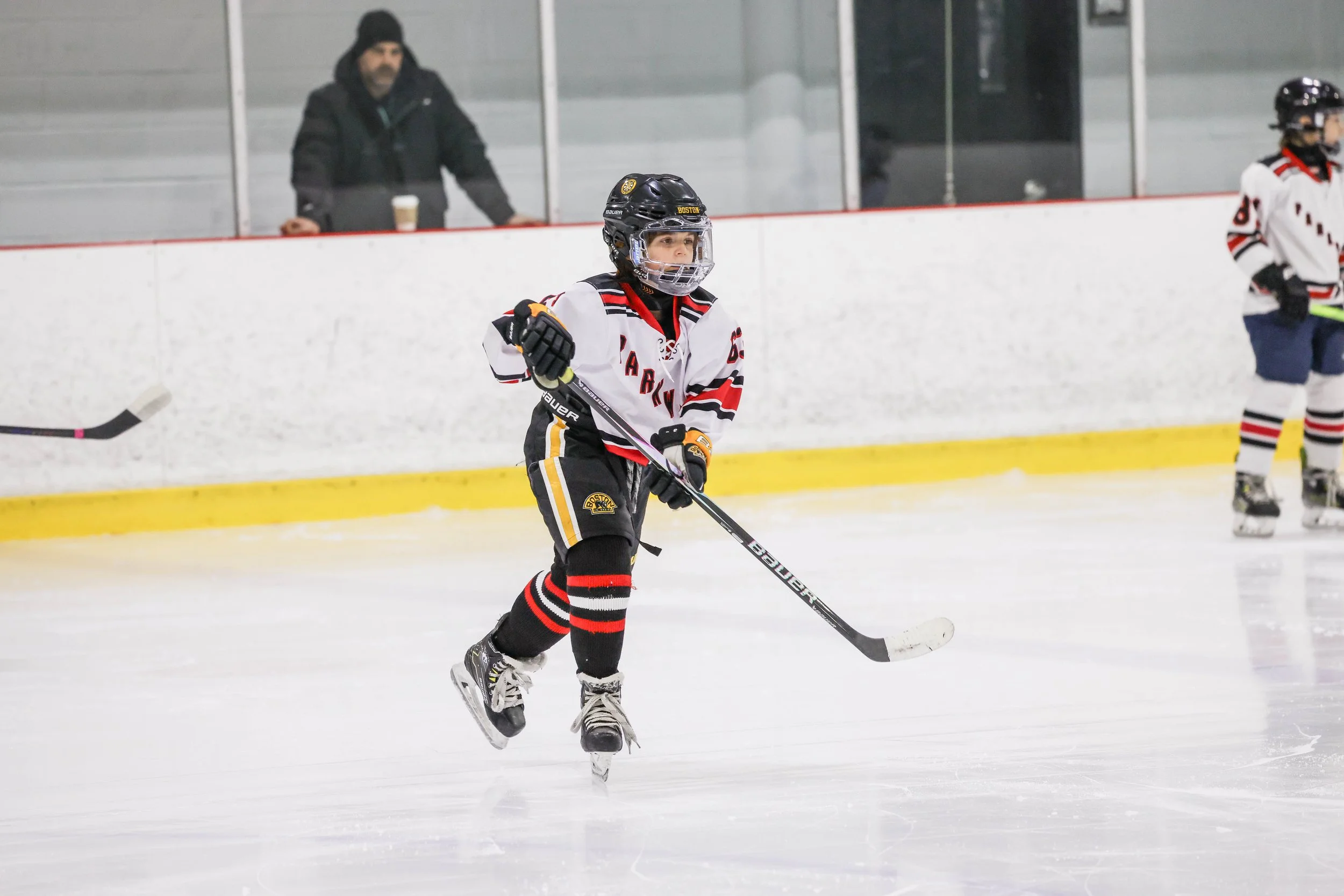Young hockey player in uniform on ice rink, holding a hockey stick, with a man watching from behind the glass in the background.