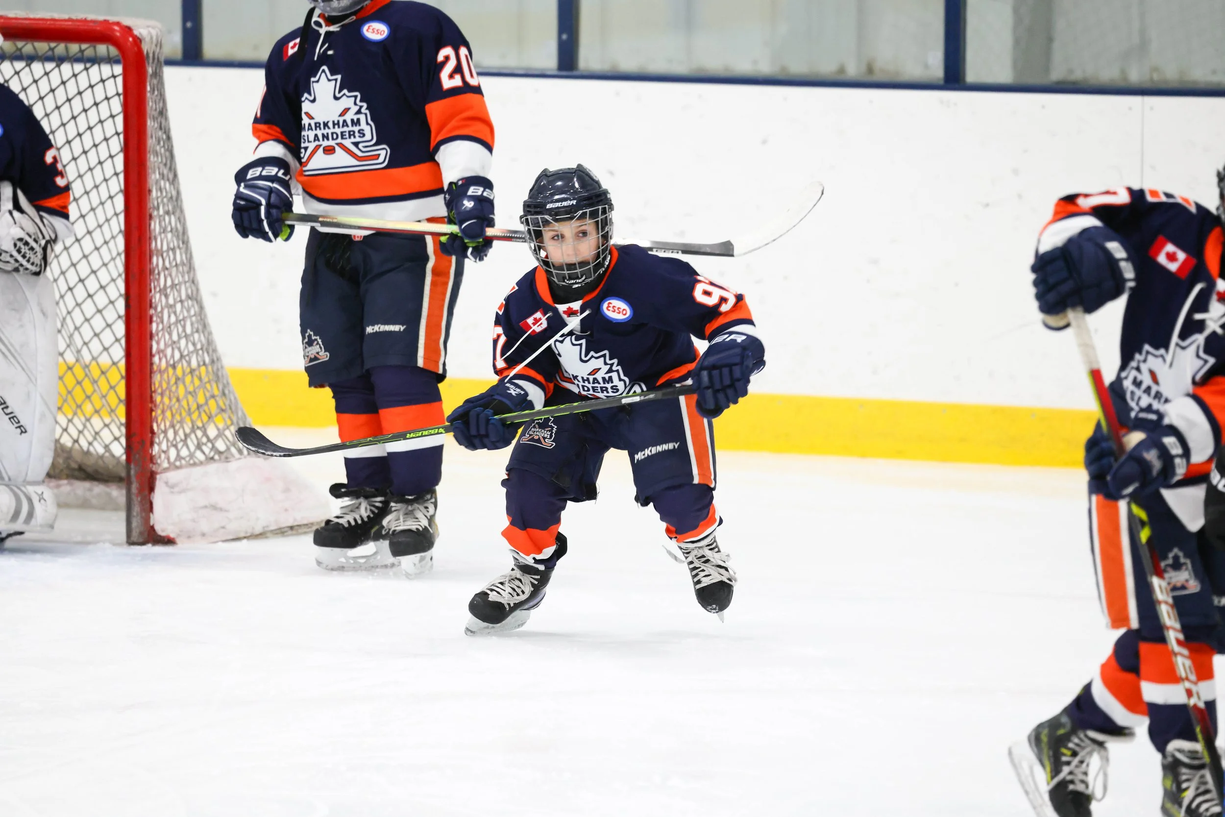 Young ice hockey player in a blue and orange uniform skates on the ice during a game, with teammates and a goalie visible in the background.