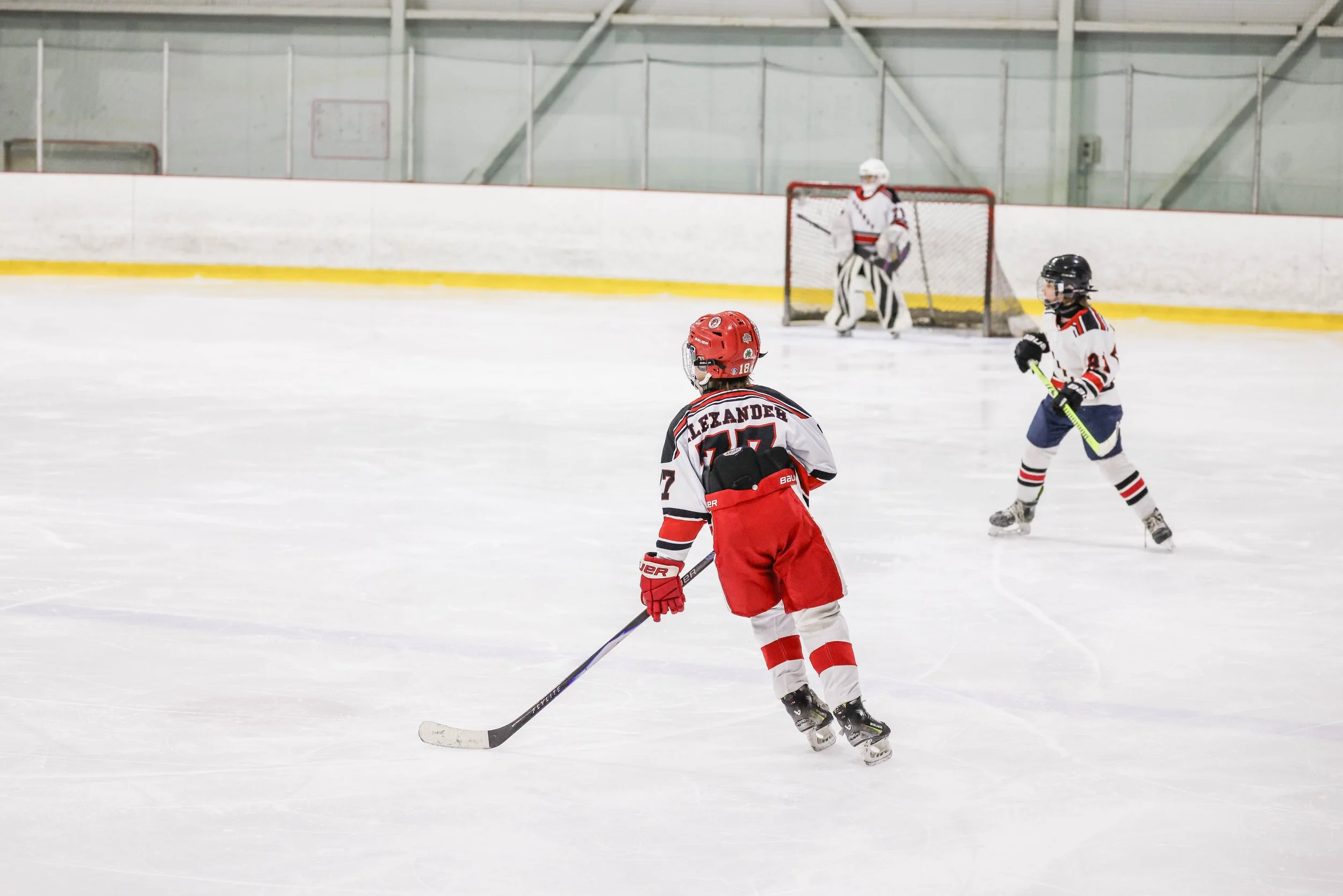 Young hockey players on the ice during a game, with one in red and white skating towards the puck, another in black and white near the goal, and a goalie in white in the background.