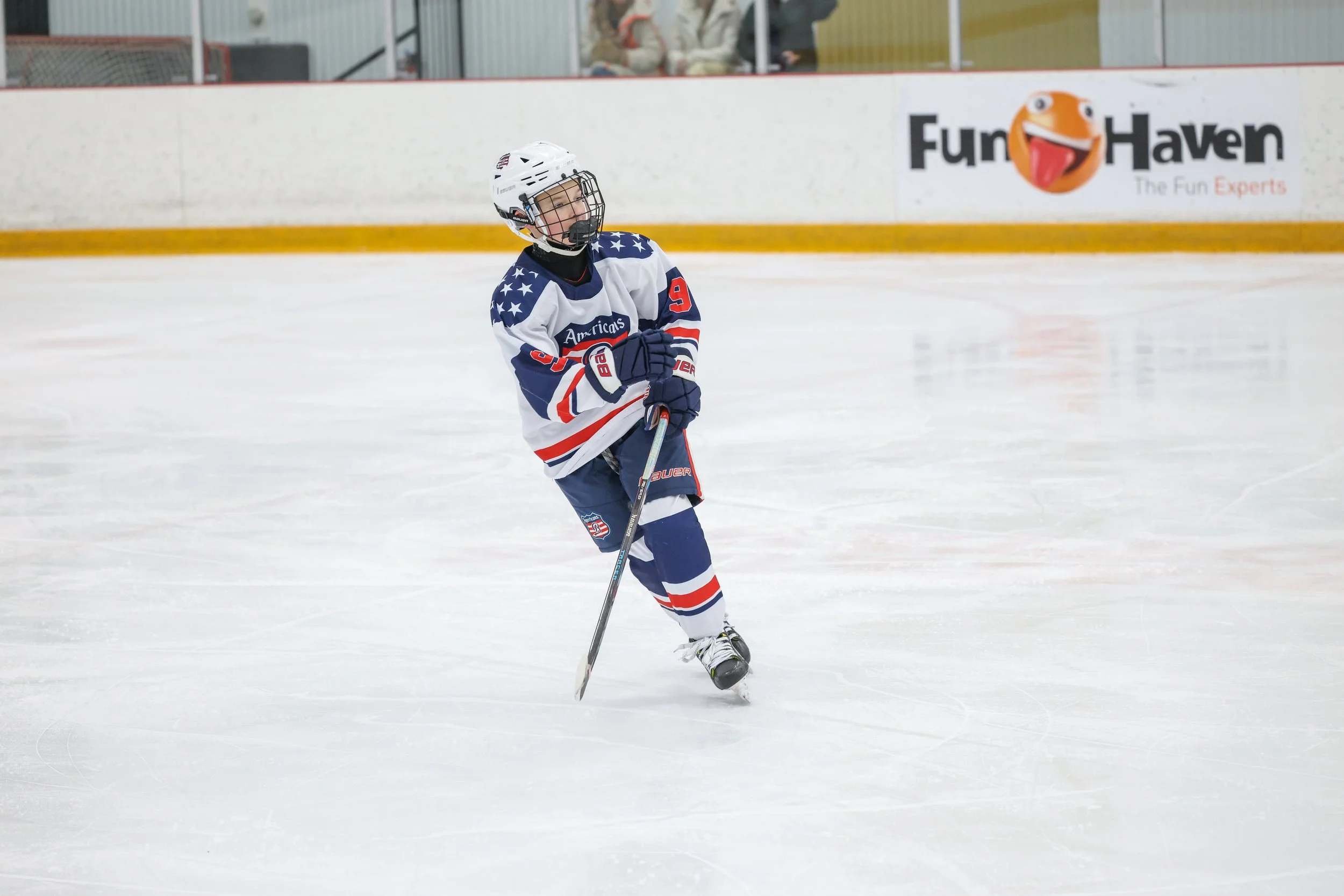 A young hockey player wearing a USA-team jersey, helmet, and gloves on the ice rink, standing with a hockey stick.