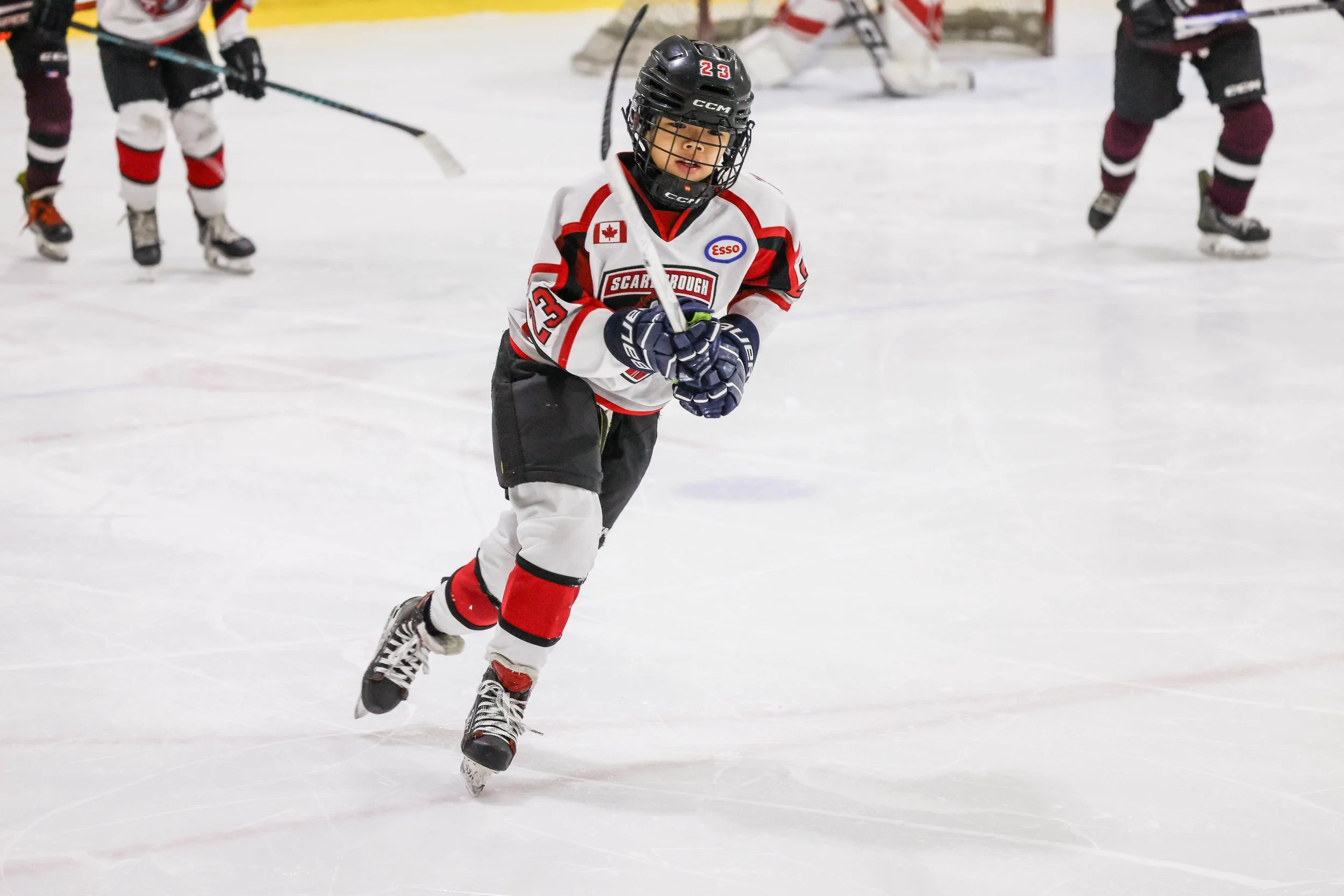 Young hockey player in a white, red, and black uniform skating on the ice rink, holding a hockey stick, with other players in the background.