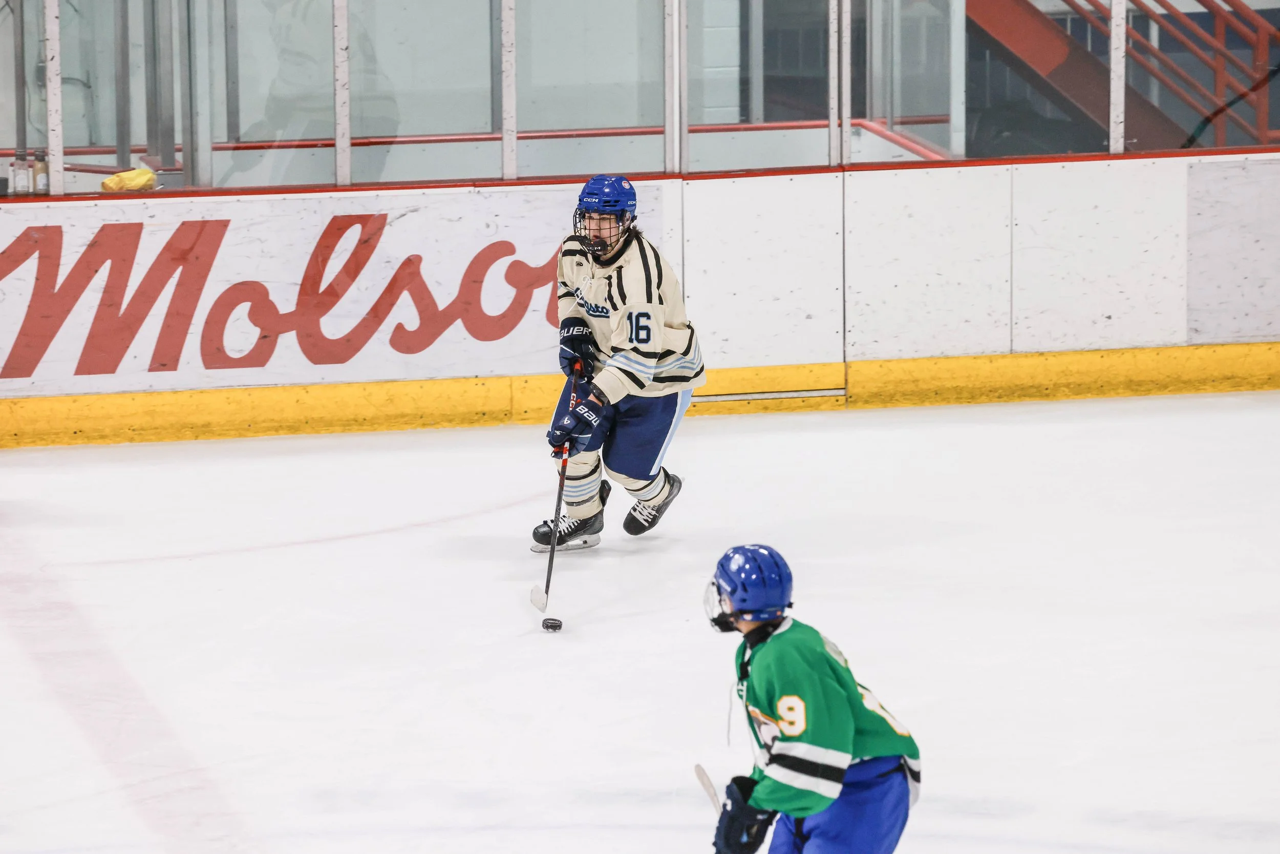 Two young hockey players on an ice rink, one in a cream and blue uniform with the number 16, wearing a blue helmet, gliding with a hockey stick, and another in a green and black uniform with the number 9, also wearing a blue helmet, in the foreground