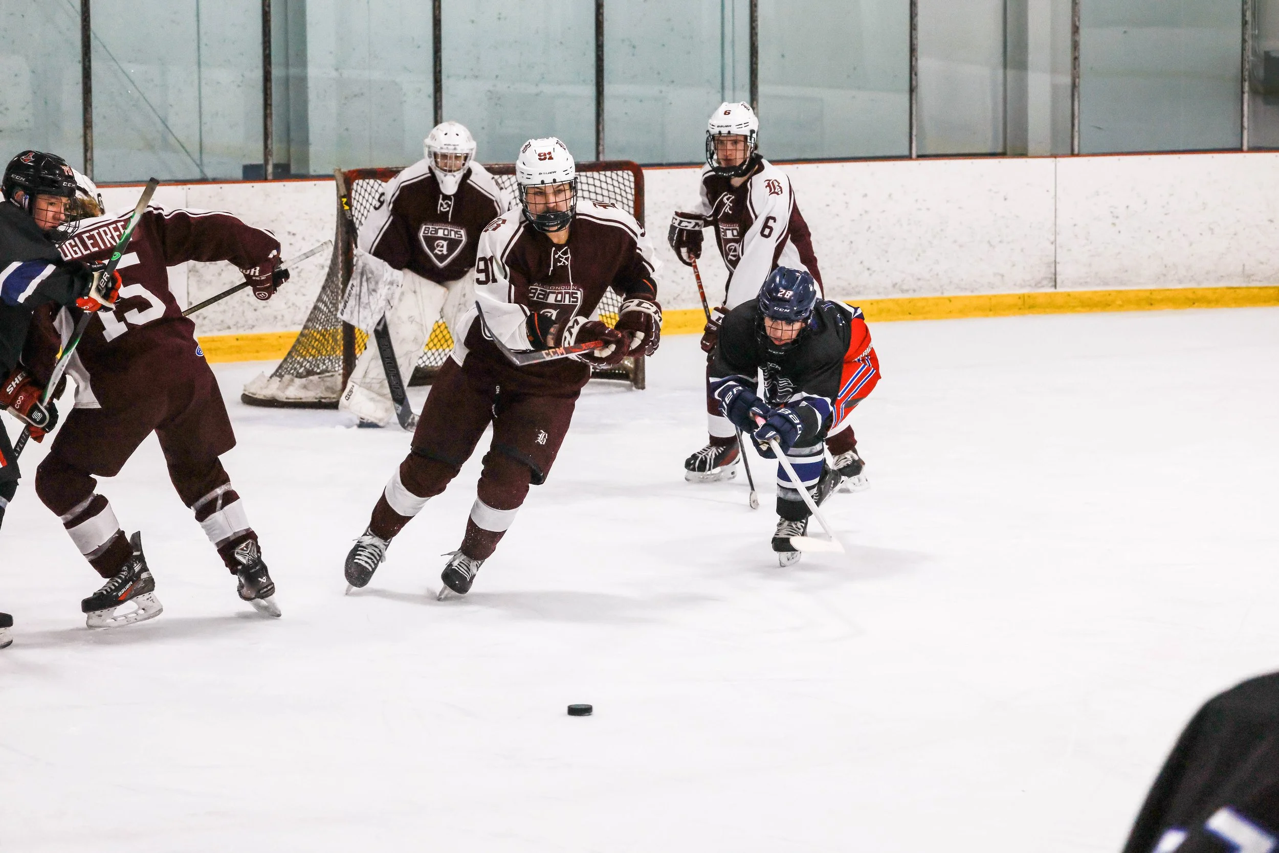 Hockey players in maroon, black, and white jerseys compete for puck on ice rink; goalie in white gear stands near goal in background.