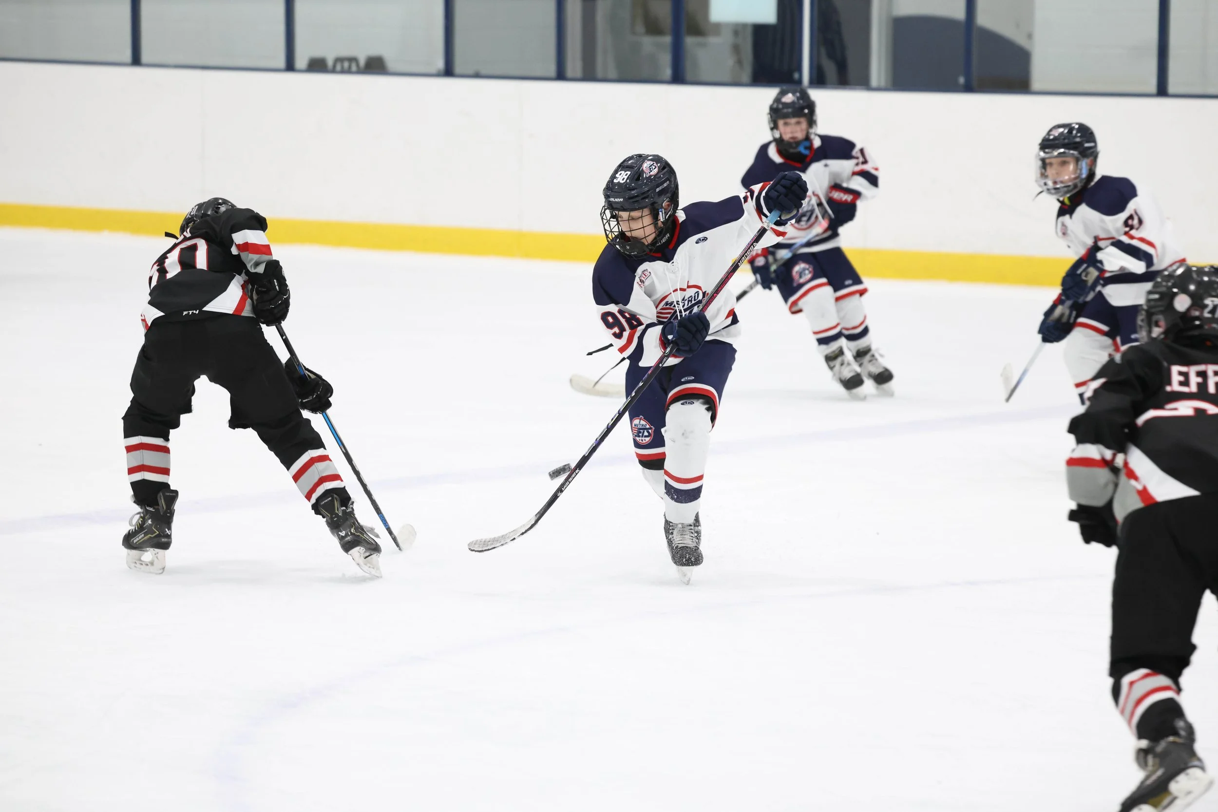 Kids playing ice hockey on an indoor rink, with one kid in a white and blue jersey about to hit the puck, surrounded by other kids in black and white jerseys.