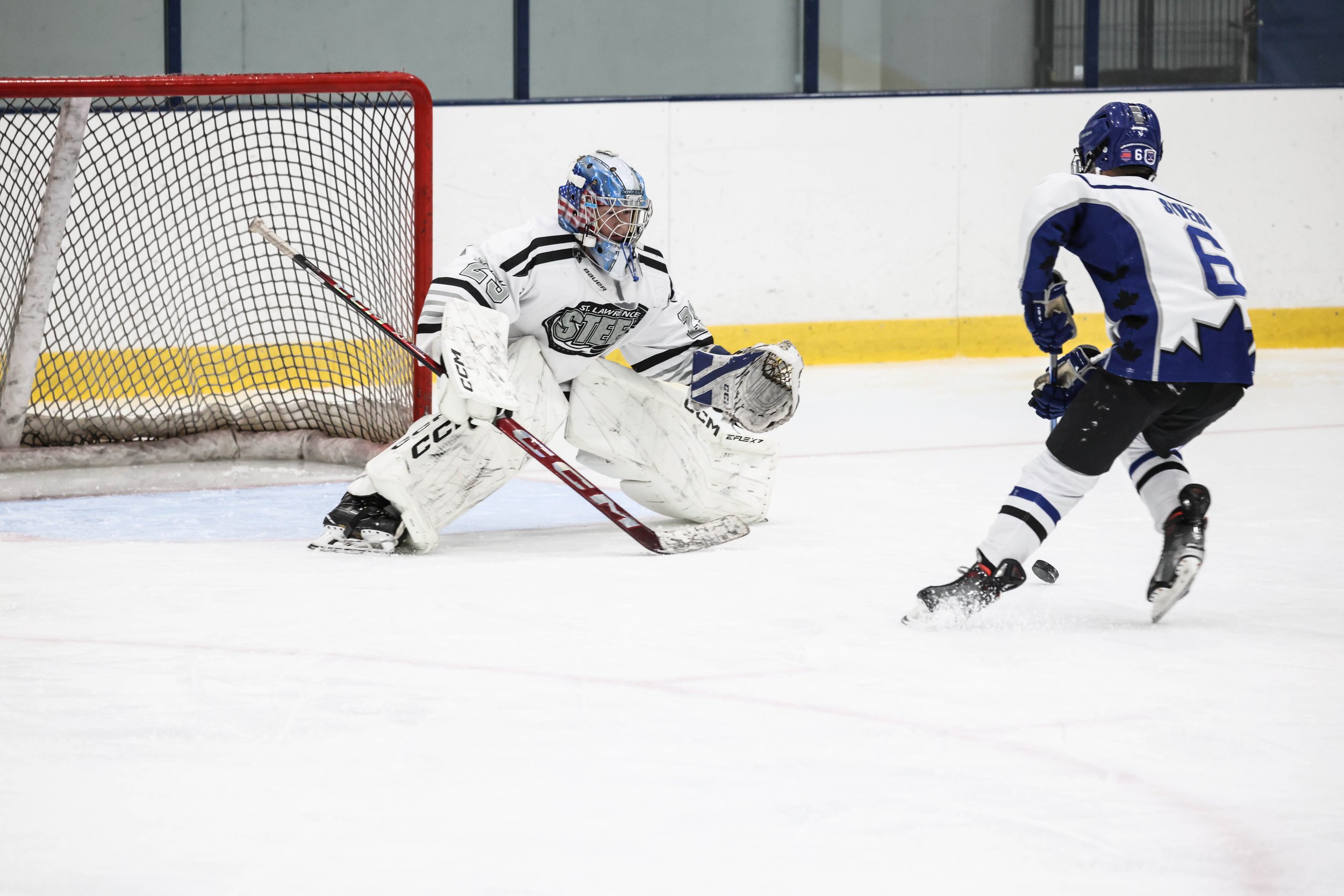 An ice hockey game showing a goalie in white gear preparing to block a shot from a player in a blue jersey near the goal.