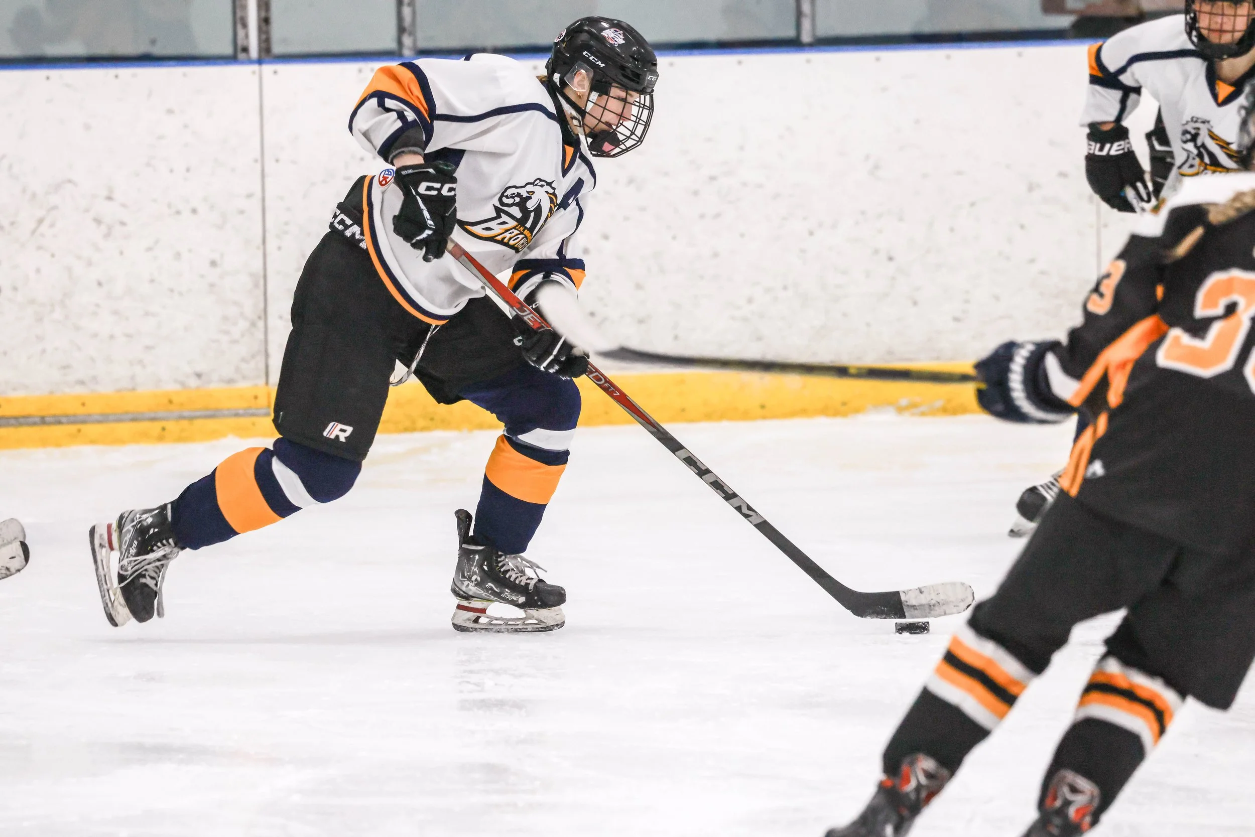 Hockey players in action on the ice, with one player in a white jersey with orange and black accents controlling the puck with his stick, while another player in a black jersey with orange and white accents reaches out.
