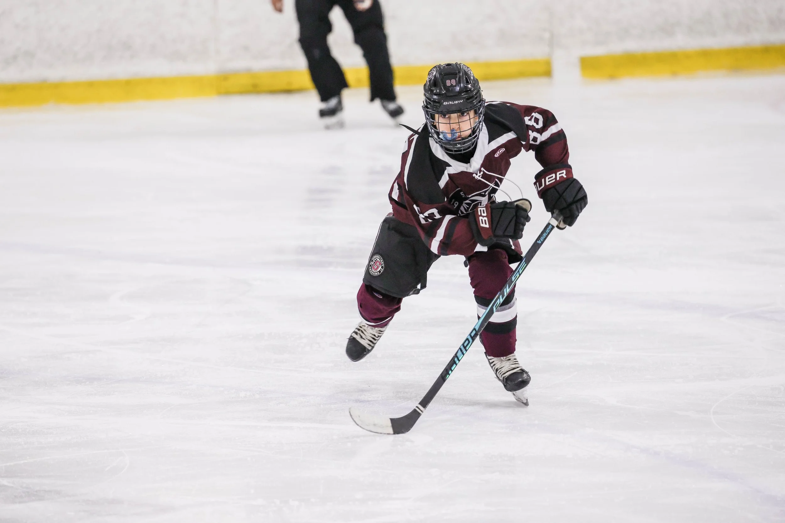 A young ice hockey player in a maroon, black, and white uniform skating on the ice with a hockey stick, wearing a helmet and gloves, with a blue mouthguard visible.