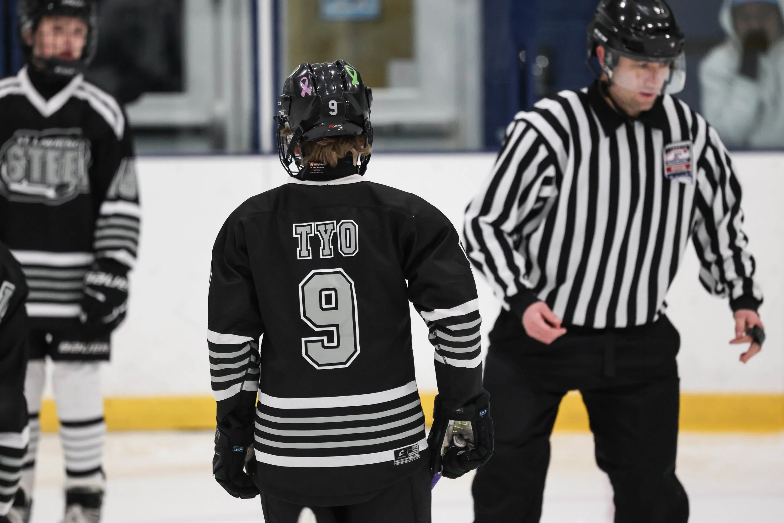 An ice hockey player with the number 9 on his black jersey, marked with the name 'TYO', standing on the ice rink facing away from the camera, engaged in a game or practice with a referee nearby.