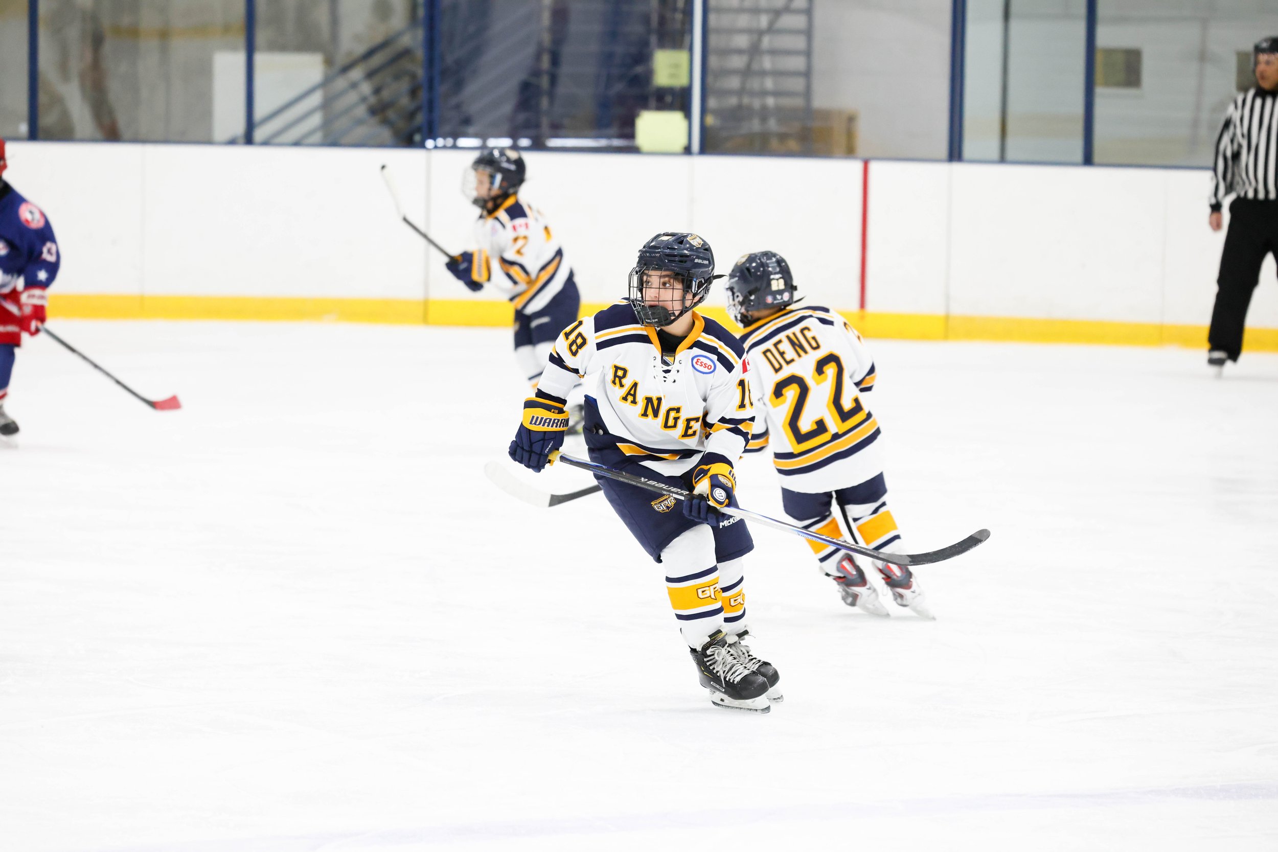 Youth hockey players in white and navy jerseys with yellow accents playing on an ice rink. One player is in the foreground, with the number 18 and the name 'RANG' on his jersey, holding a hockey stick. Behind him, another player with the number 22 an