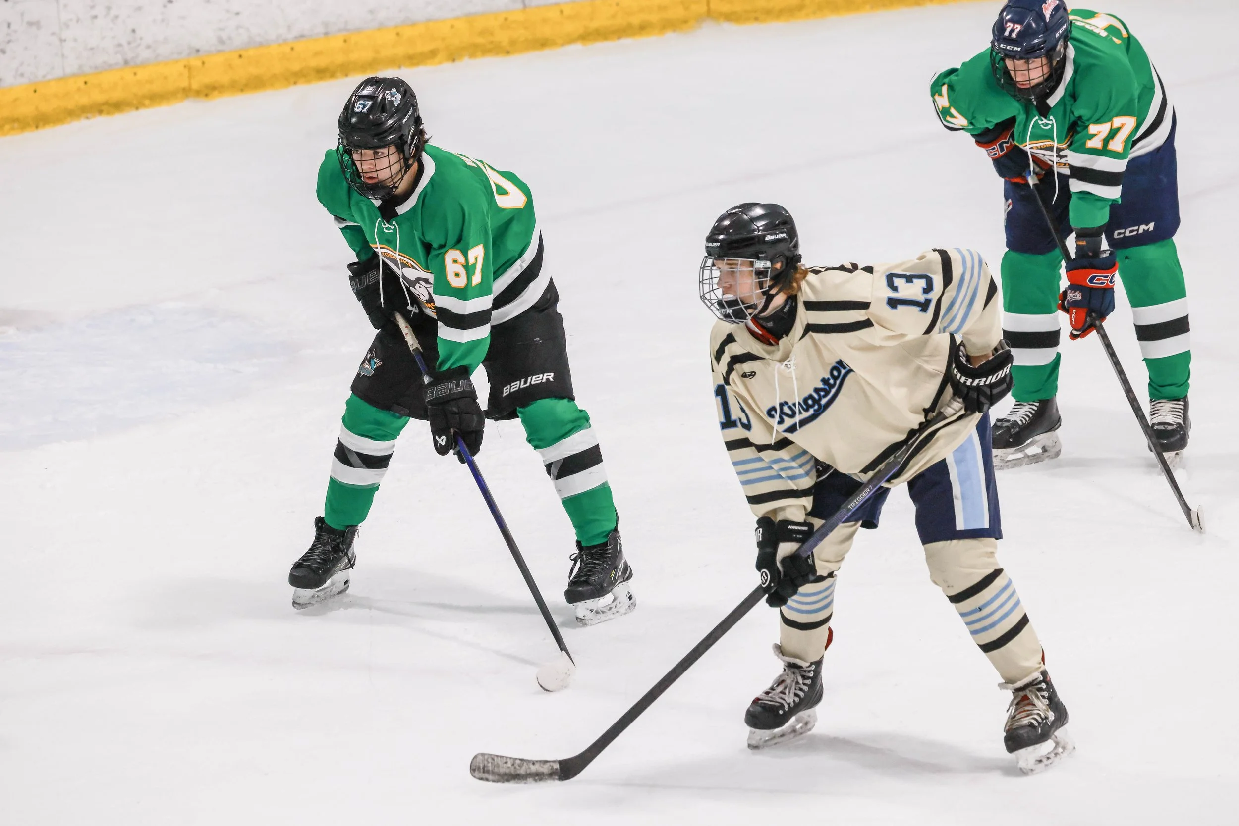 Three hockey players on ice during a game, two in green jerseys and one in a beige jersey, competing for control of the puck.