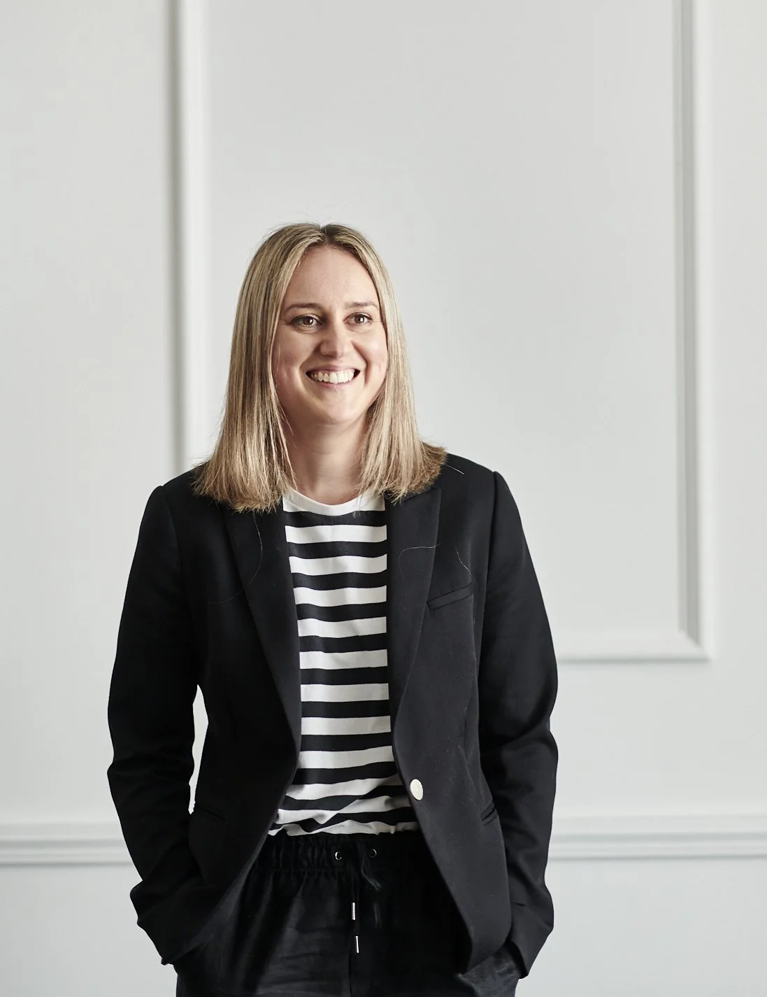 A woman smiling and looking to the side, wearing a black blazer and a black-and-white striped shirt, standing in a room with light-colored walls and decorative paneling.
