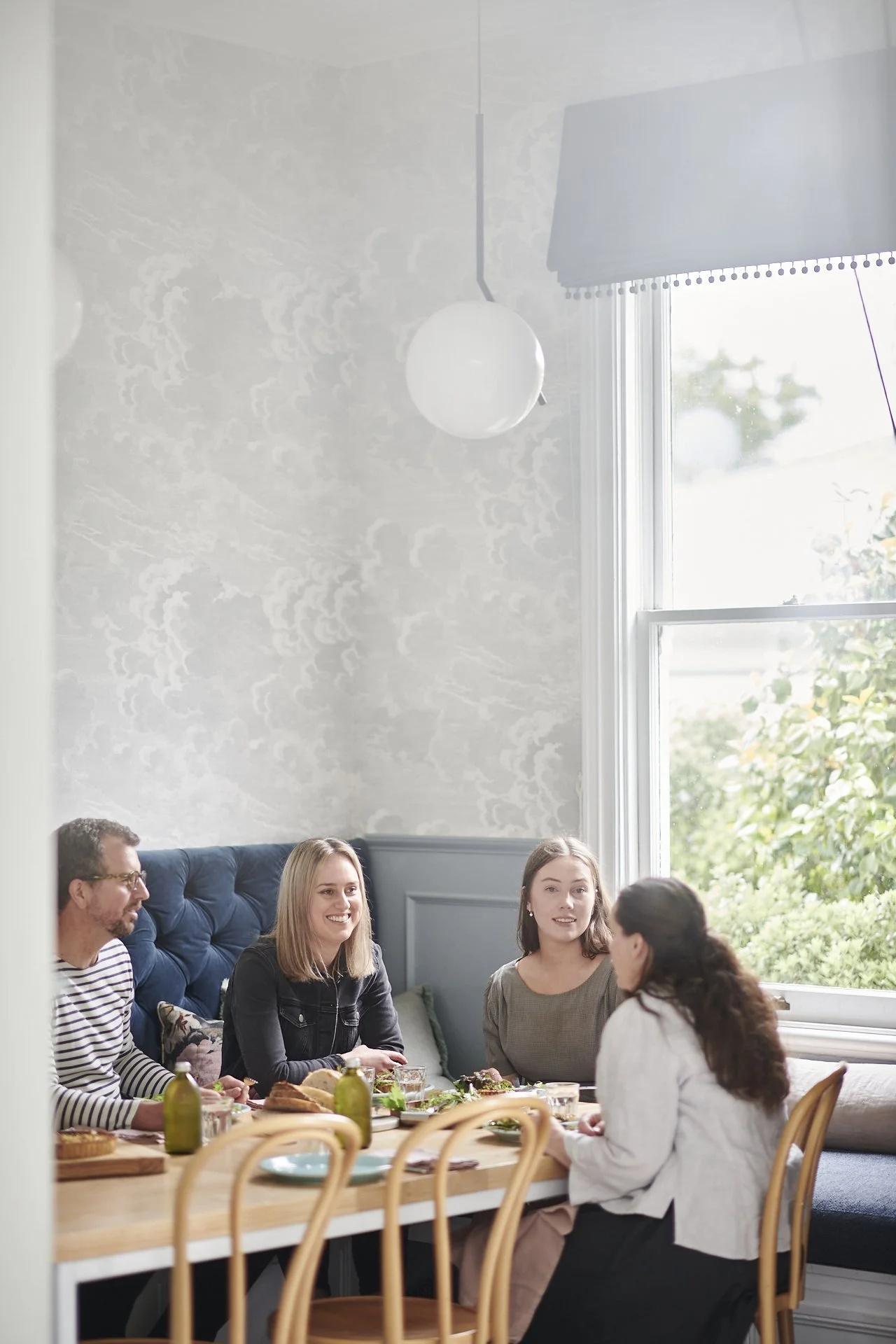 Four people sitting around a dining table, engaged in conversation and enjoying a meal, with a window showing greenery outside.