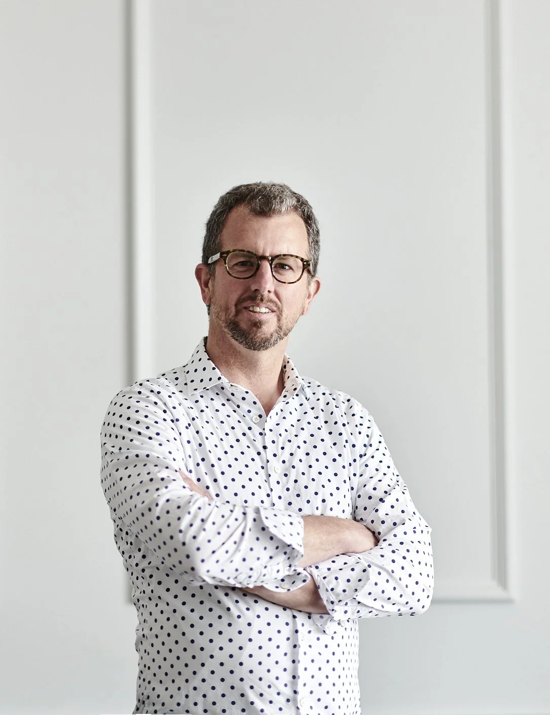Man with glasses and a beard wearing a white shirt with black polka dots, standing with arms crossed in front of a plain wall.