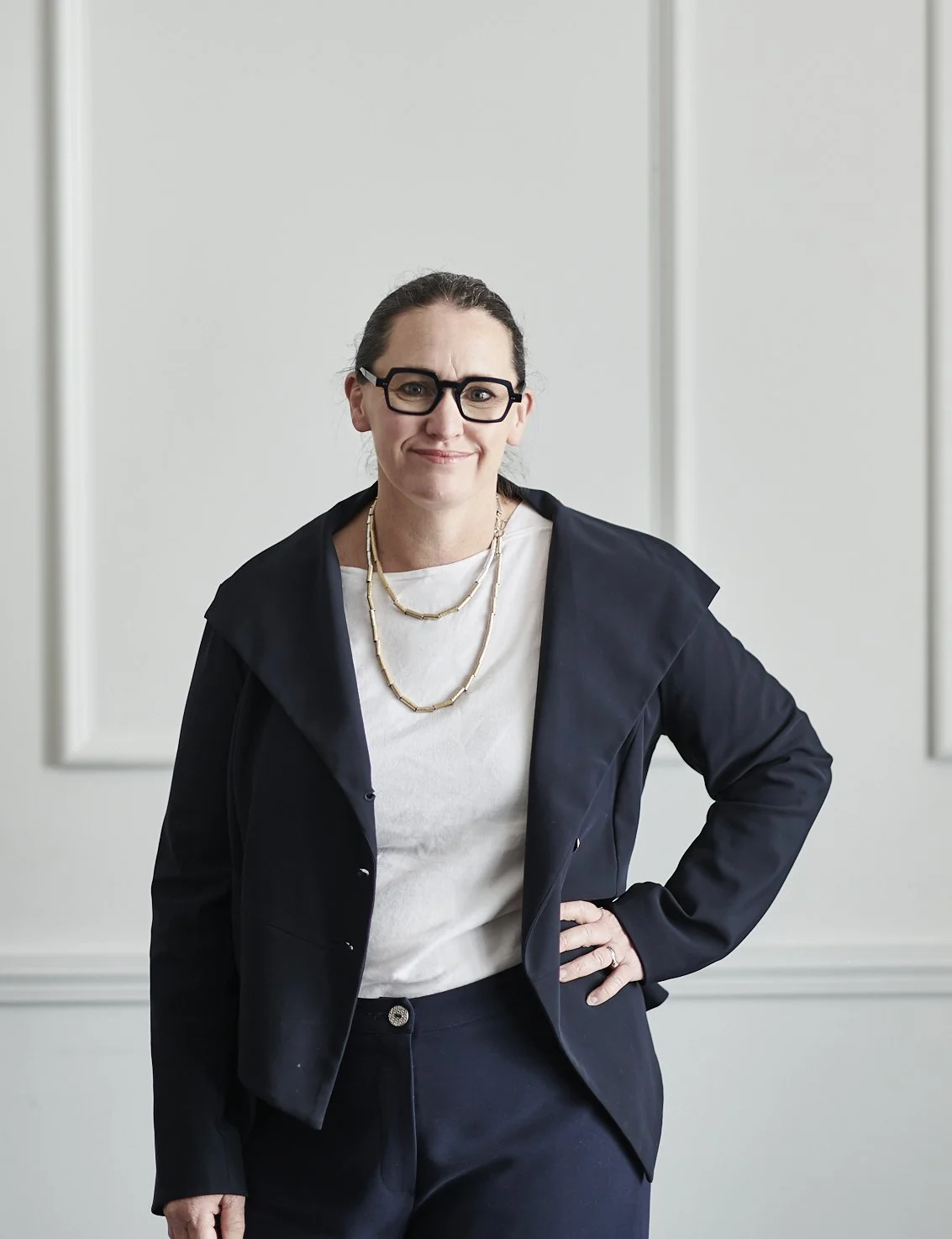 A woman with dark hair tied back, wearing glasses, a black blazer, a white top, and layered necklaces, standing with her hand on her hip against a light-colored wall.