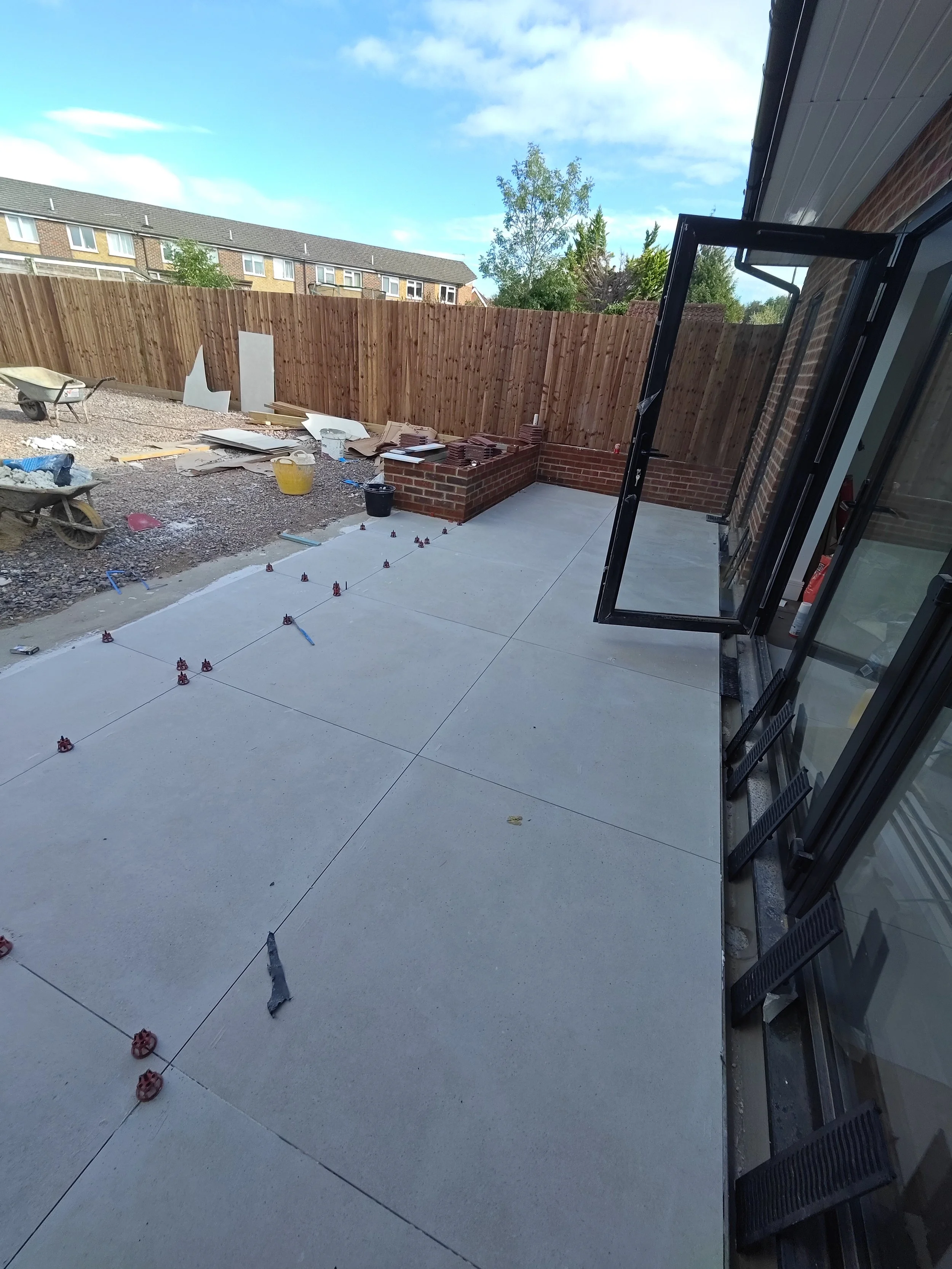 A backyard under construction with a new concrete patio, construction materials, and tools. The yard is enclosed with a wooden fence, and there is a brick wall with stacked bricks. An open window or door frame is visible on the right side.