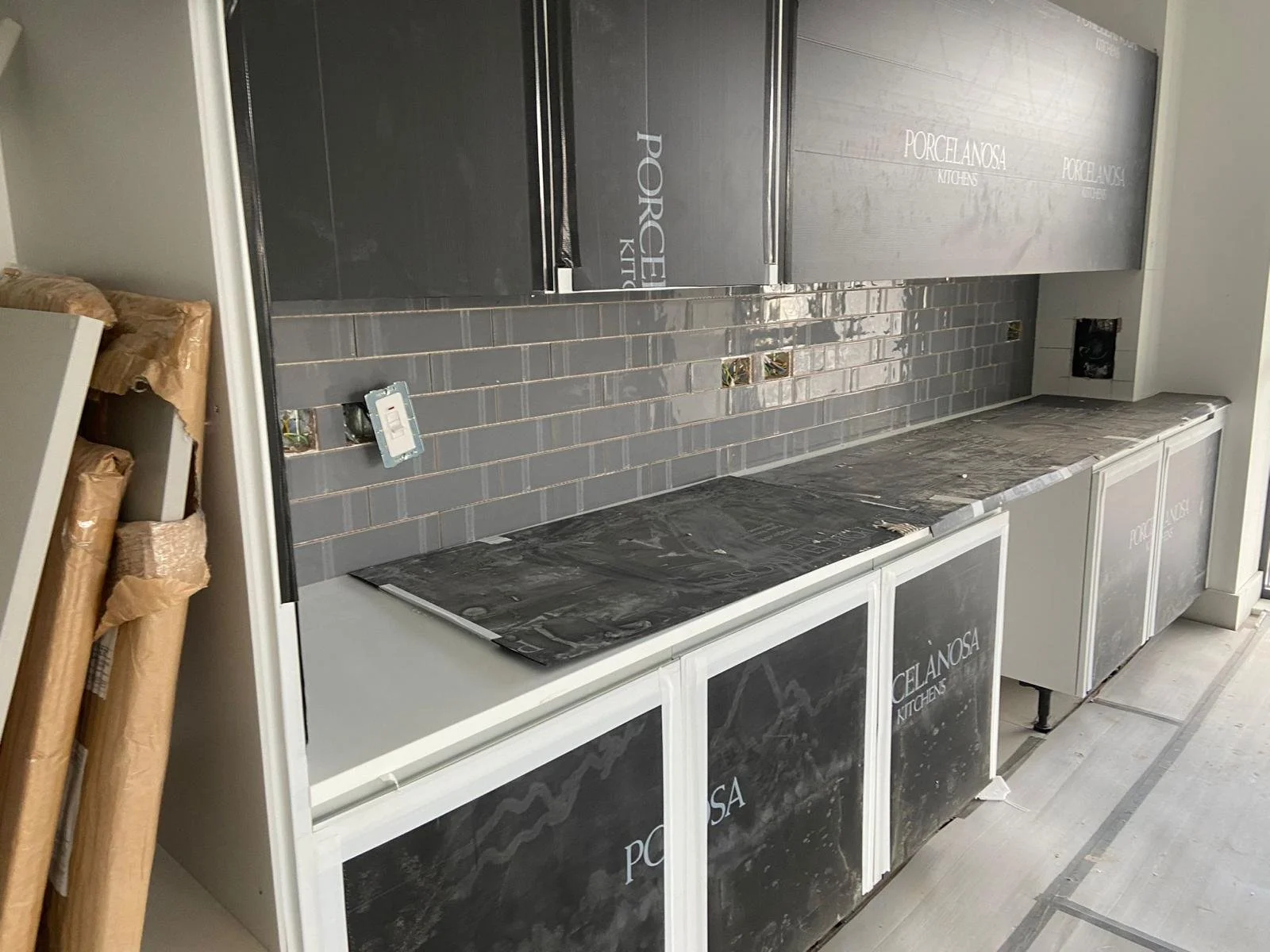 Under-construction kitchen with gray cabinets, gray subway tile backsplash, and black countertop, with some packaging materials on the left side.
