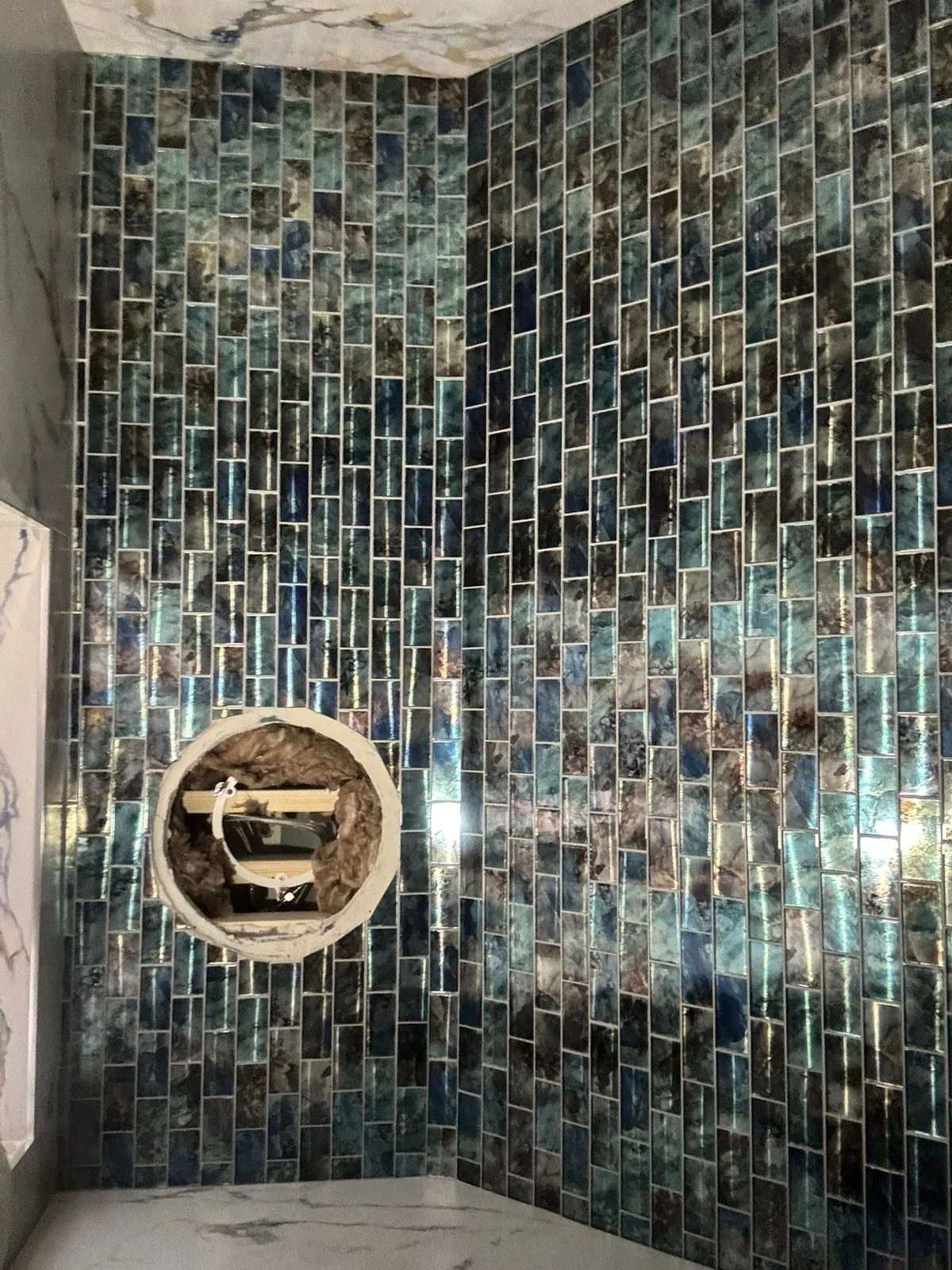 Bathroom wall with iridescent mosaic tiles and a round cutout for a mirror or medicine cabinet, containing construction materials.