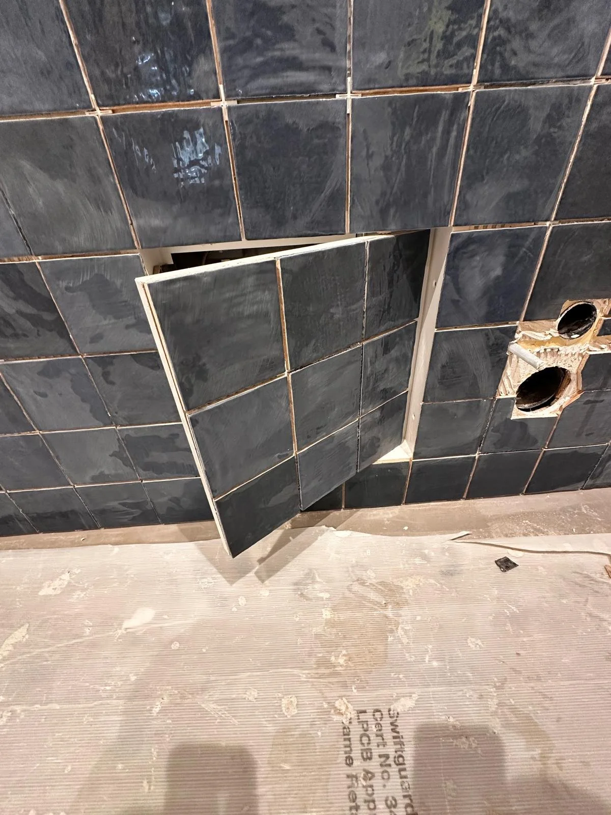 Black tiled wall with two holes and a partially removed tile, with construction materials and a wooden surface at the bottom.
