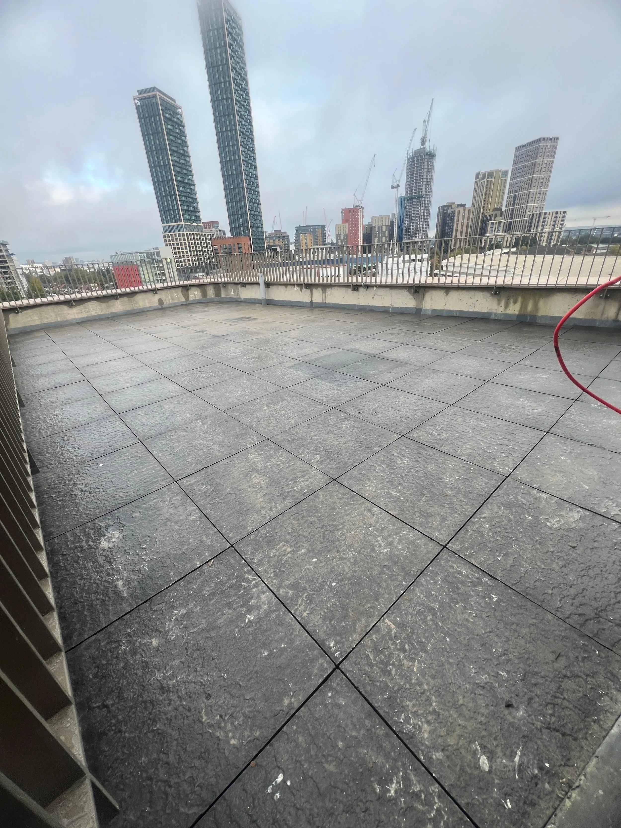 A rooftop with gray tiled flooring, metal railing, and a view of tall modern buildings under a cloudy sky.