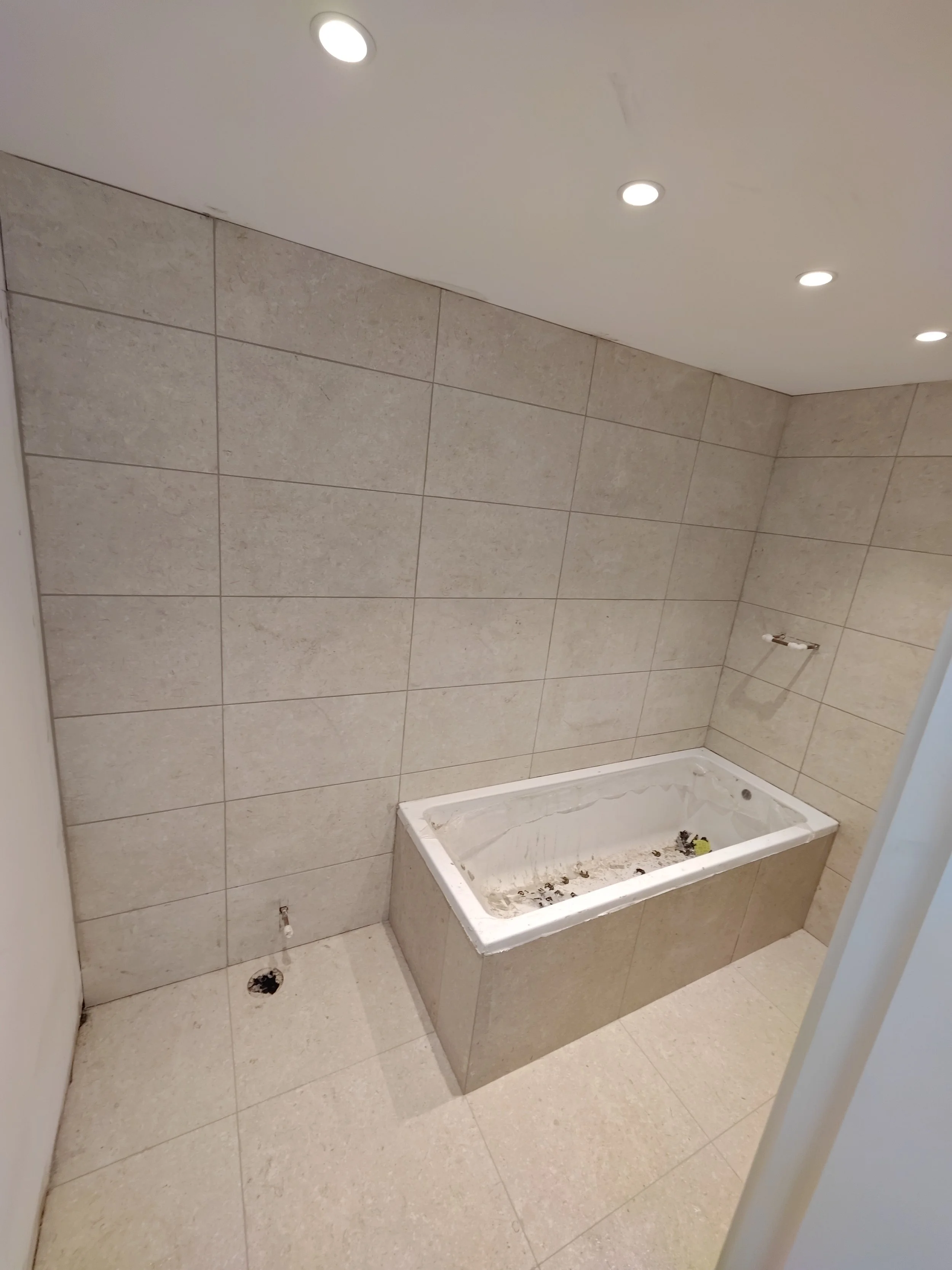 An unfinished bathroom with large beige tile walls and a partially installed bathtub with debris inside. Recessed ceiling lights are visible, and there is an electrical outlet without a cover on the wall.