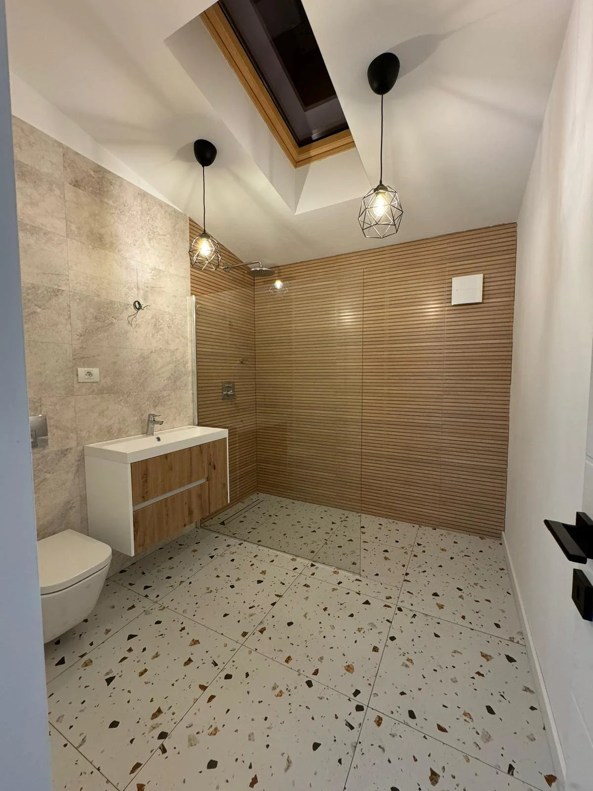 Modern bathroom with terrazzo floor, beige and wood wall tiles, a small white sink with wooden cabinet, and a walk-in shower area with wood slat wall, skylight, and hanging pendant lights.