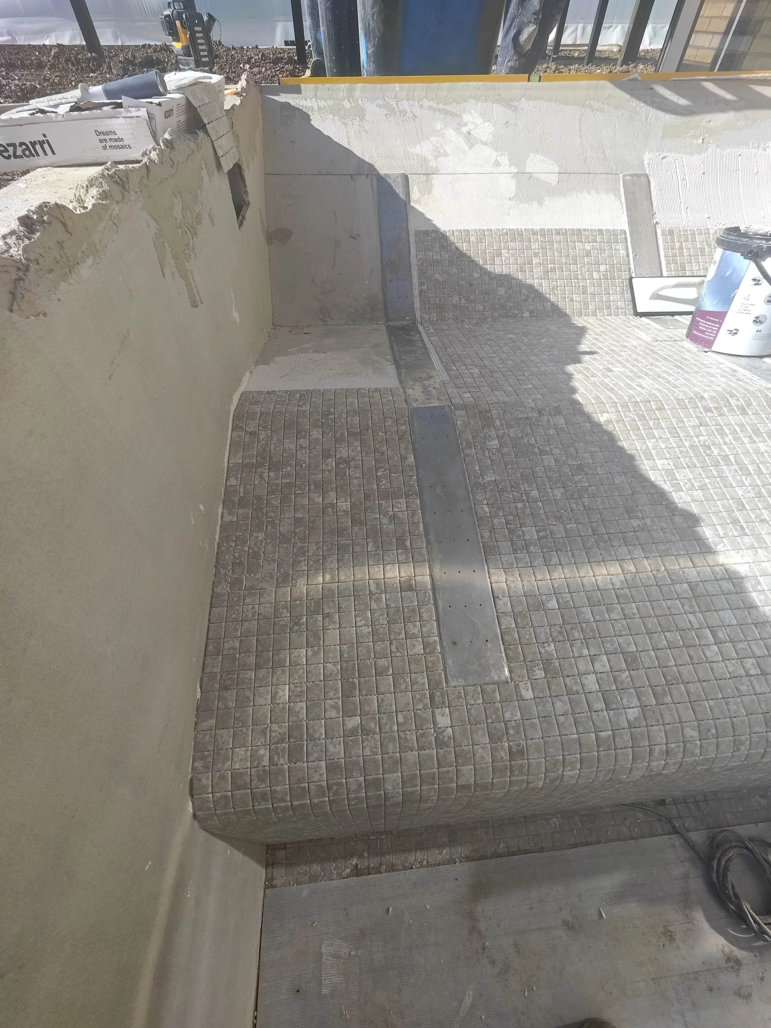 Construction site showing a partially finished wall, tiled floors, a bucket, and construction tools. Bright sunlight causes shadows on the wall and floor.