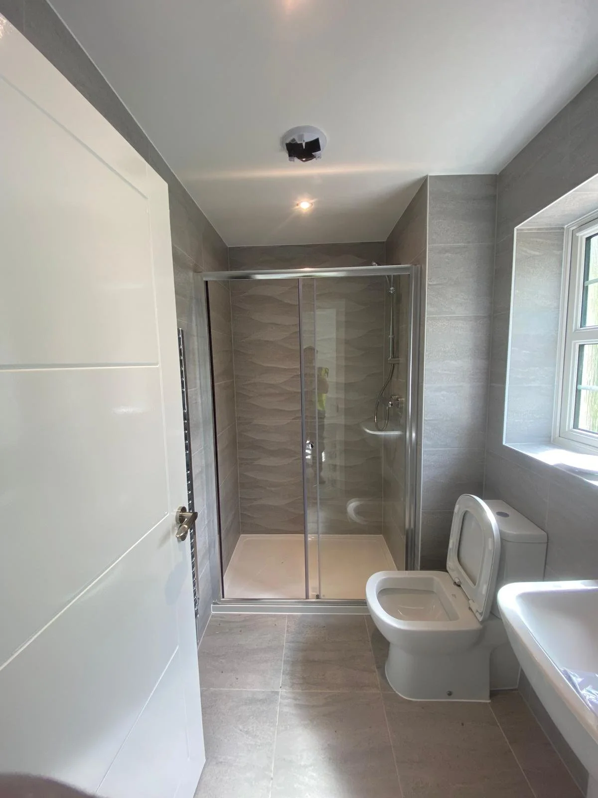 Modern bathroom with a glass shower enclosure, a white toilet with the lid open, a window letting in natural light, and gray tiled walls and floor.