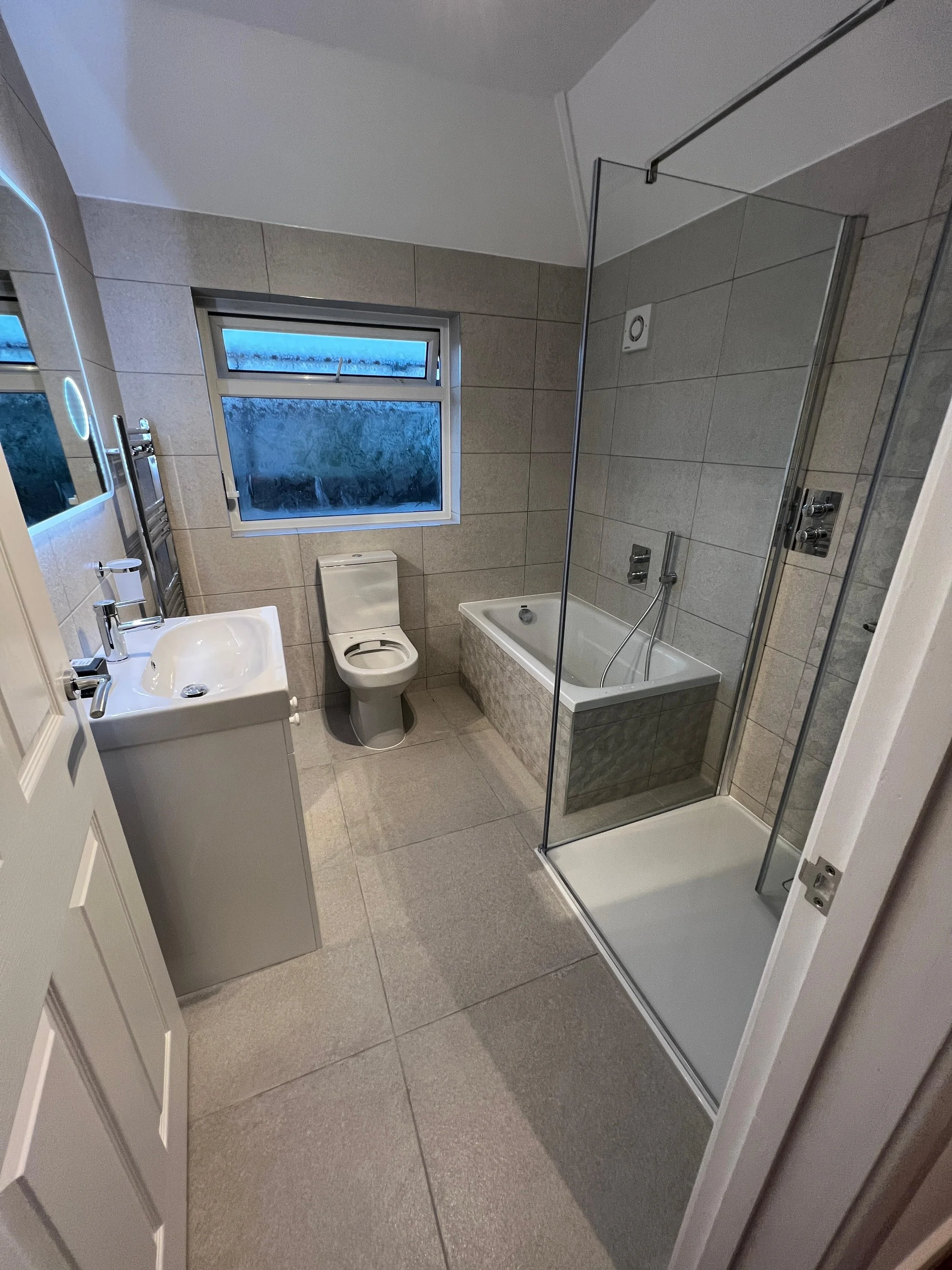 Bathroom with beige tiled floor and walls, a white sink with a faucet on the left, a small toilet in the center, and a bathtub with a shower curtain on the right. There is a window with frosted glass above the toilet, and a shower stall with a glass door on the far right.