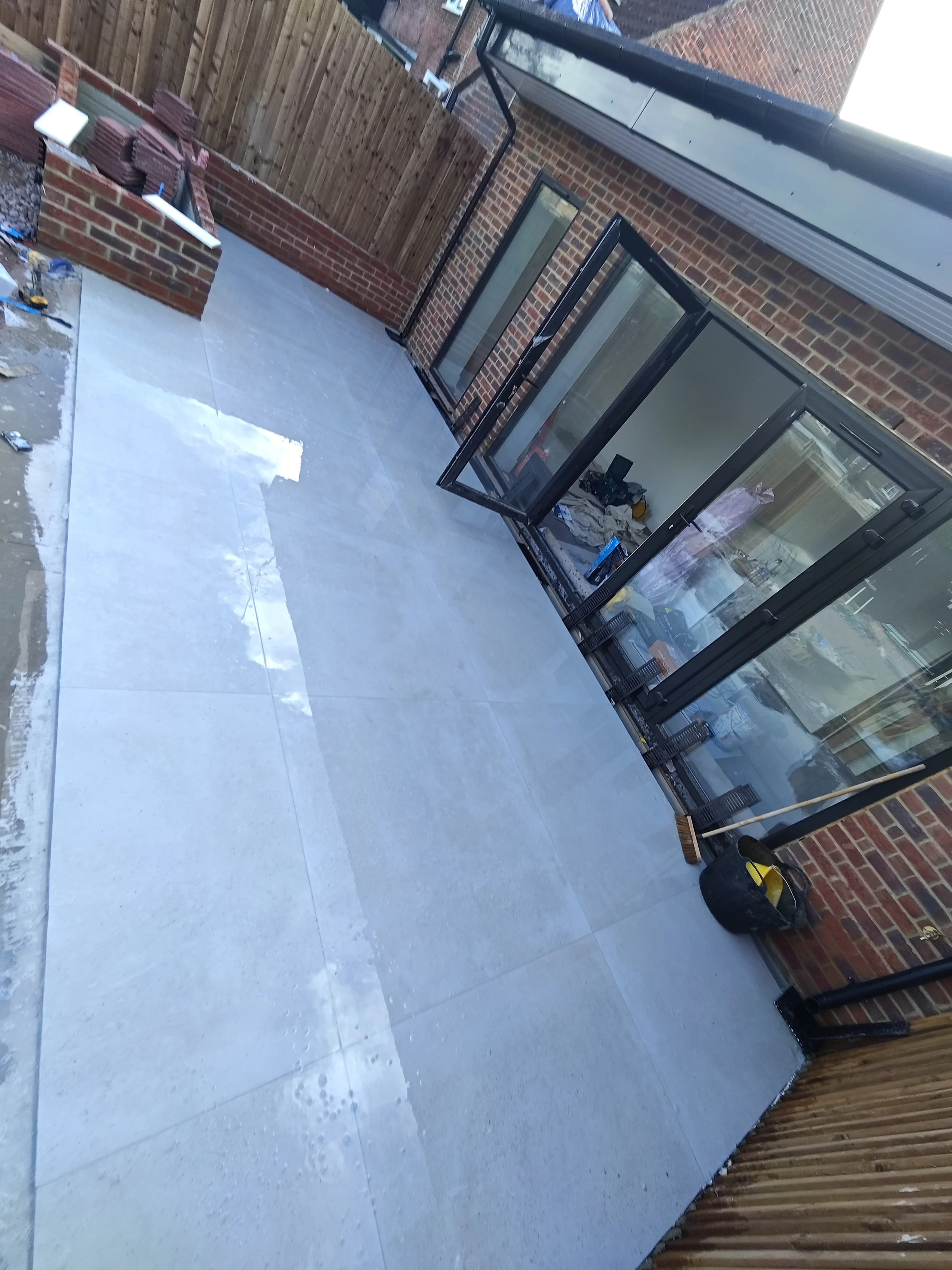An outdoor patio area with a newly installed light gray tiled floor, a brick wall with a black-framed glass enclosure, a wooden fence, and various construction tools and materials scattered around.