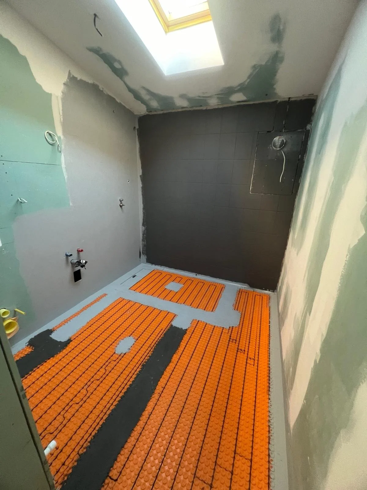 A bathroom under construction with orange radiant floor heating mats installed, gray walls, and a ceiling with a skylight.