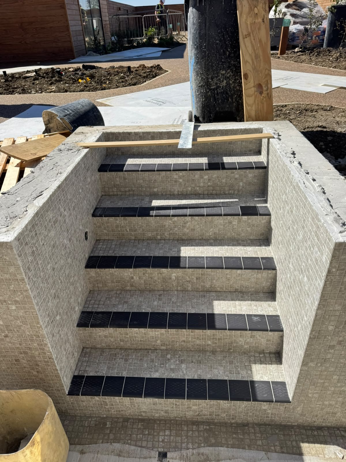 Under construction of a small tiled pool with black tile accents on steps, surrounded by construction materials and dirt, with workers and building in background.
