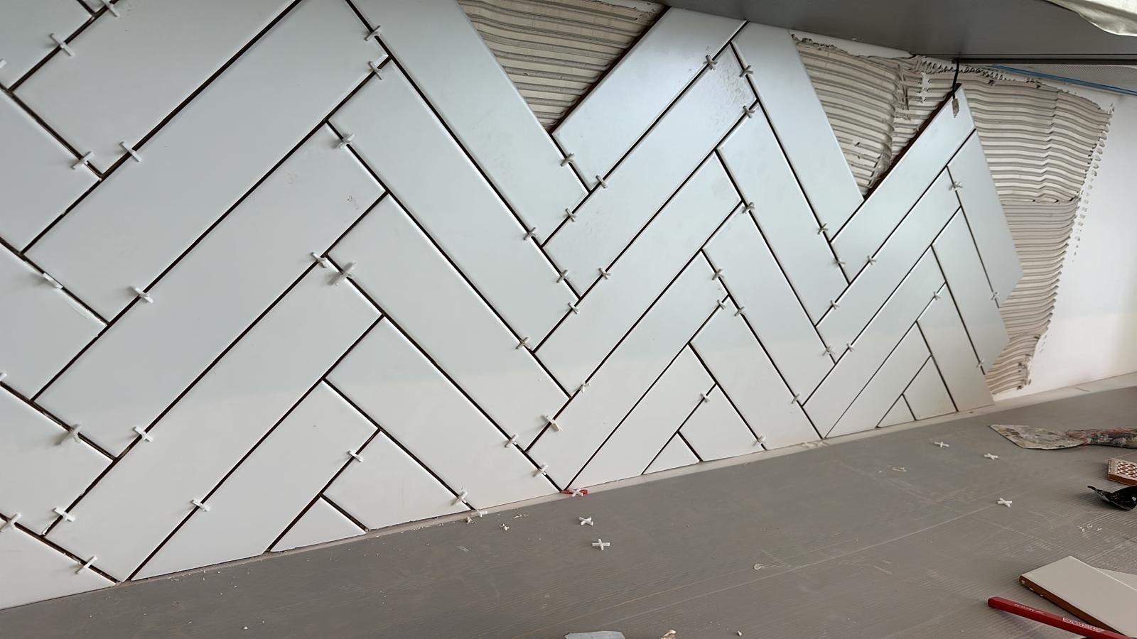 Interior wall with white rectangular tiles arranged in a herringbone pattern under construction, with thin black spacers visible between tiles and some construction tools and materials on the floor.