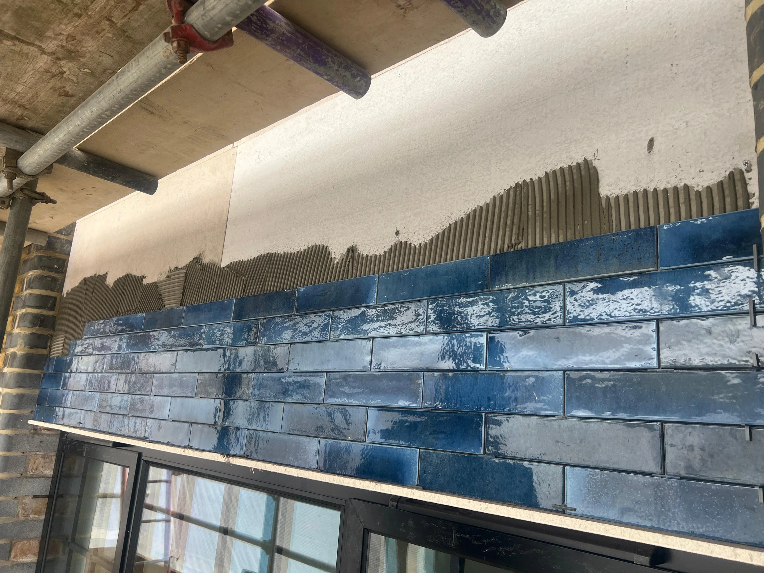 Interior view of a building under construction, showing a wall with partially installed blue ceramic tiles, window frames, and construction scaffolding.