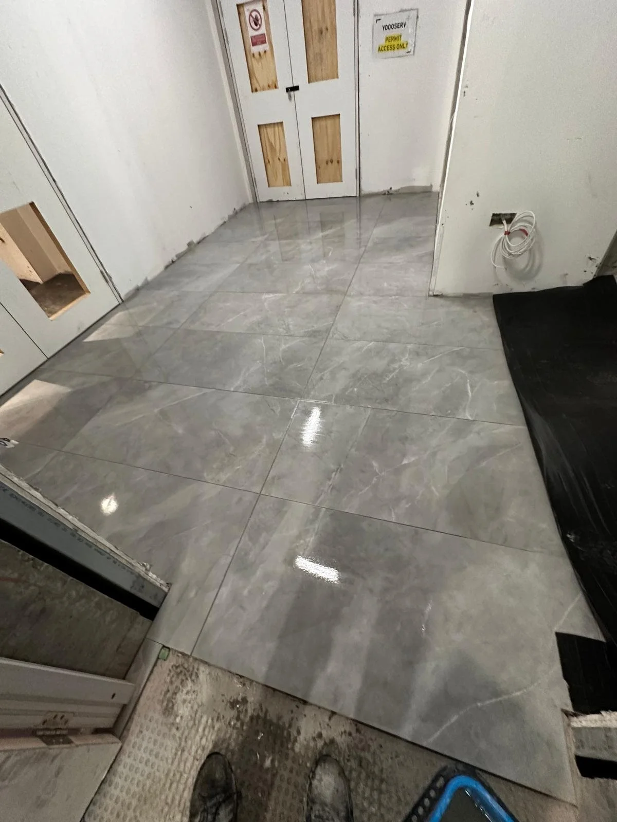 Newly tiled shiny gray marble floor in an unfinished room with white walls, a door with wood panels, and a black protective covering on one side.