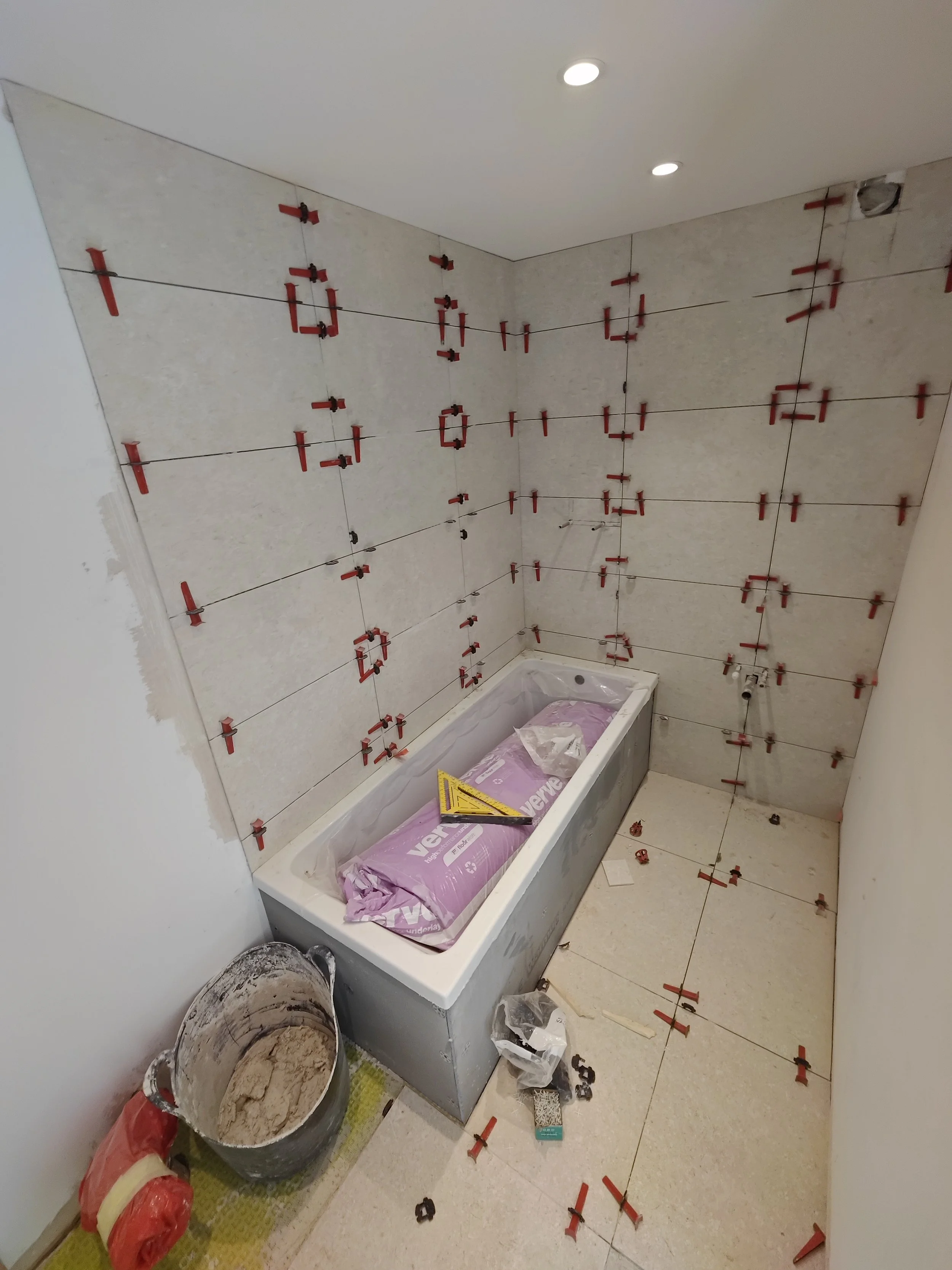 A bathroom under construction with tiled walls, a bathtub covered with a pink protective sheet, construction tools, and materials scattered around.