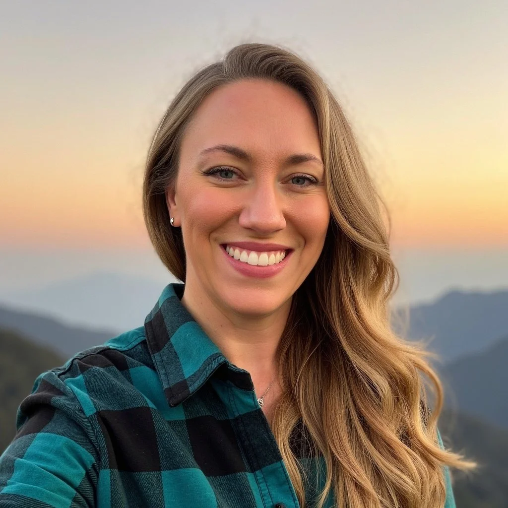 A woman with long wavy hair smiling outdoors during sunset with mountains and a colorful sky in the background.