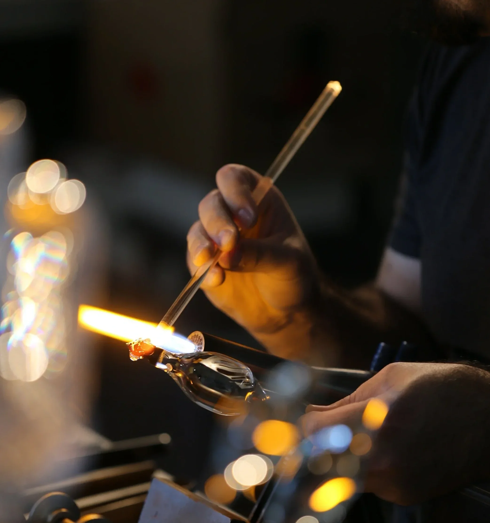 A glassblower shaping molten glass with a long metal rod in a dark studio.