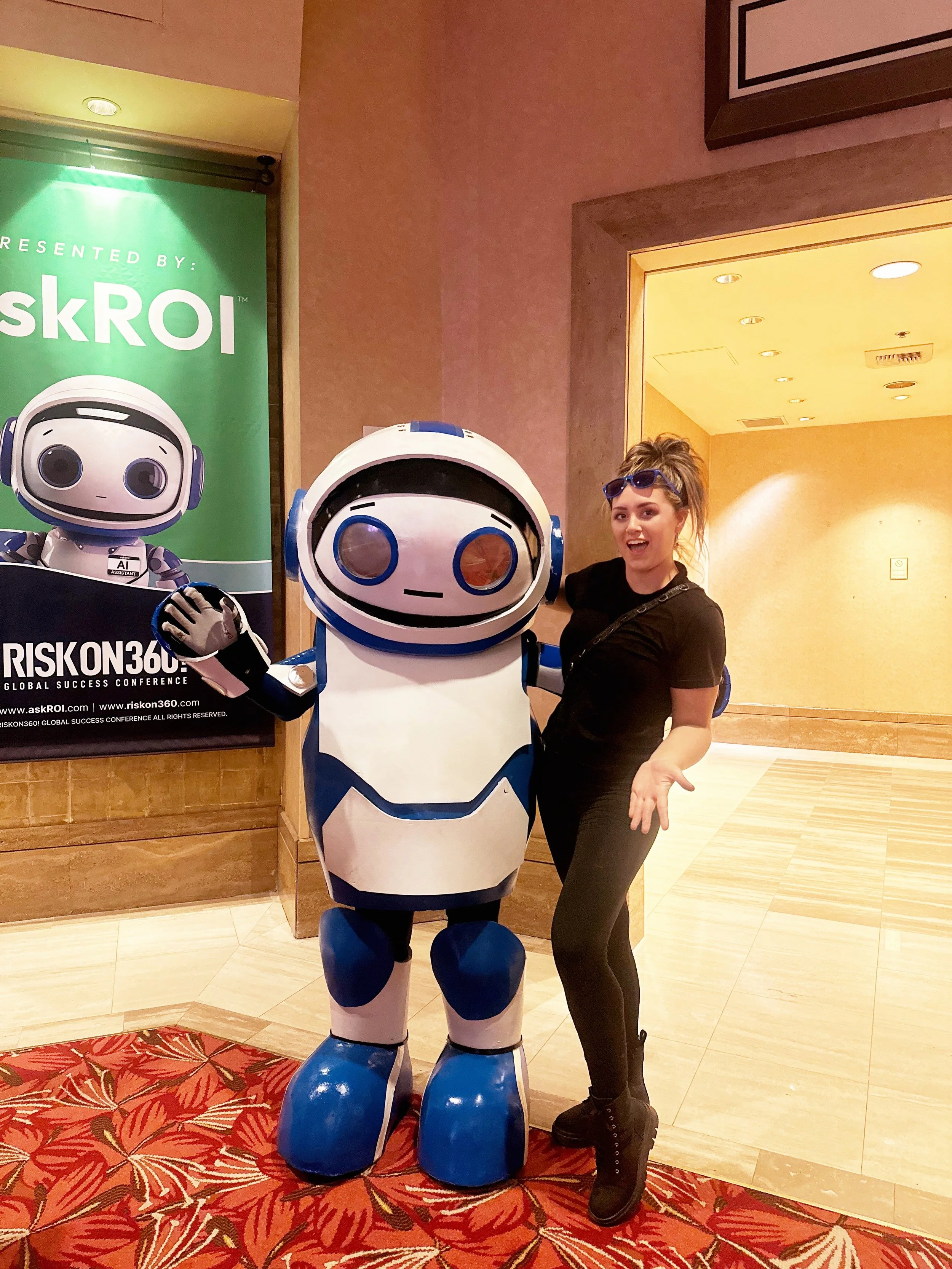 A woman in black clothing with sunglasses on her head standing next to a large blue and white robot mascot at a conference. The woman is smiling and gesturing with her hands toward the robot.