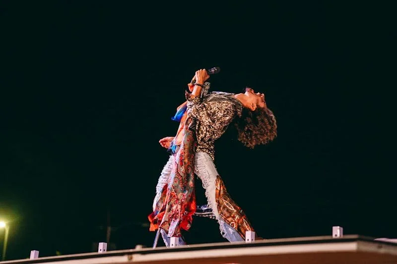 Performer singing on stage at night, with head thrown back and microphone in hand, dressed in patterned and colorful clothing.