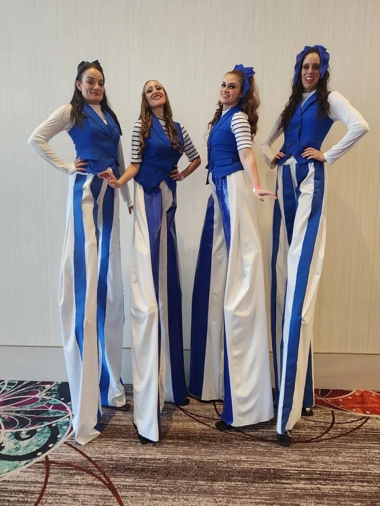 Four women dressed as tall, vintage-style, striped performers, standing indoors against a plain wall, smiling and posing for the photo.