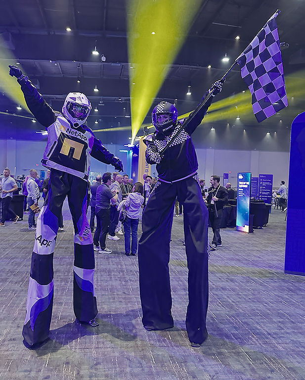 Two people on stilts dressed in racing-themed costumes at an indoor event. One in a racing suit with a helmet and racing numbers, the other in black with checkered flag, both striking celebratory poses.