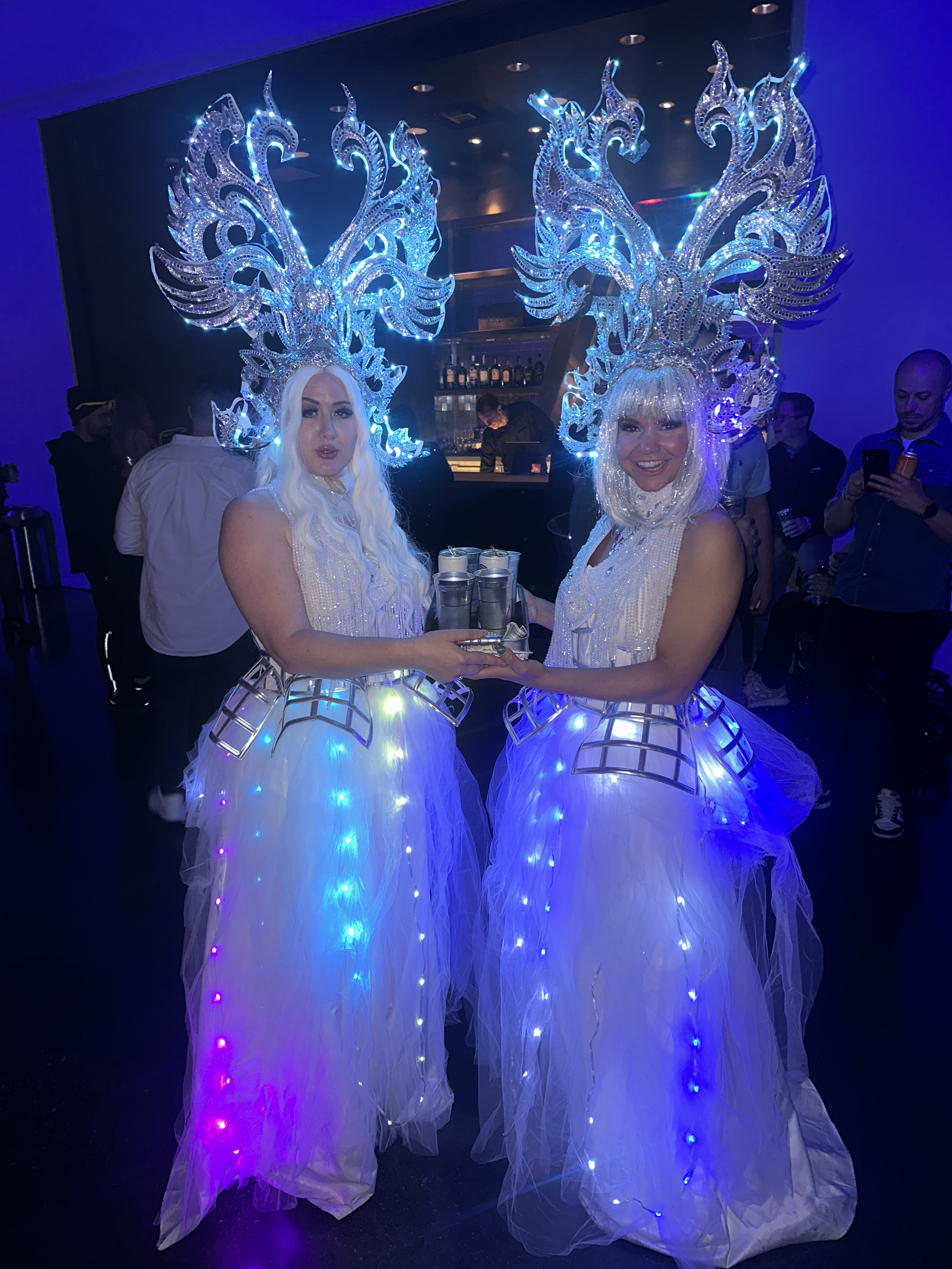 Two women dressed in elaborate white costumes with large illuminated headdresses, holding a tray of drinks at a party or event.