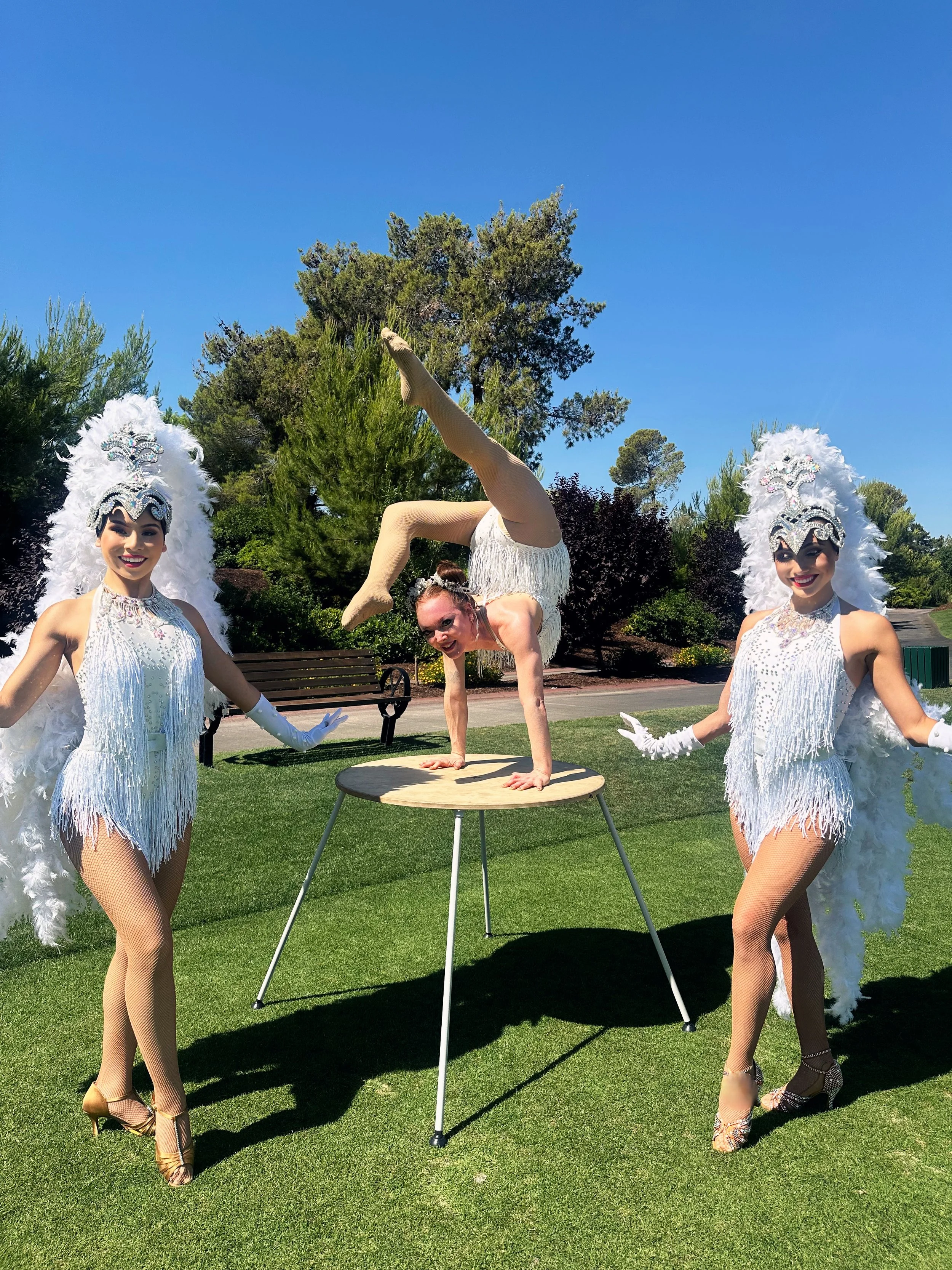 A performer doing a handstand on a round table, surrounded by two women dressed as showgirls with feathered headdresses and costumes, outdoors on a sunny day with trees in the background.
