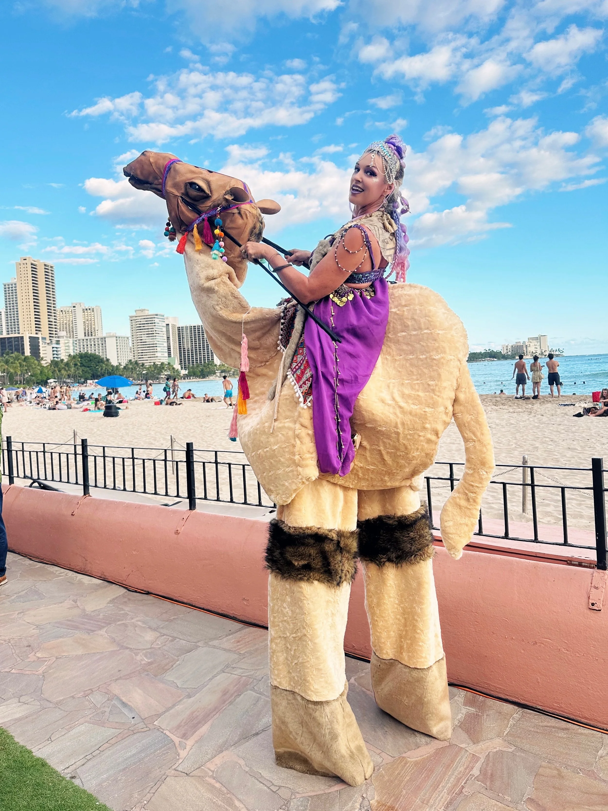 A woman dressed in a colorful costume riding a camel on the beach with city buildings in the background.