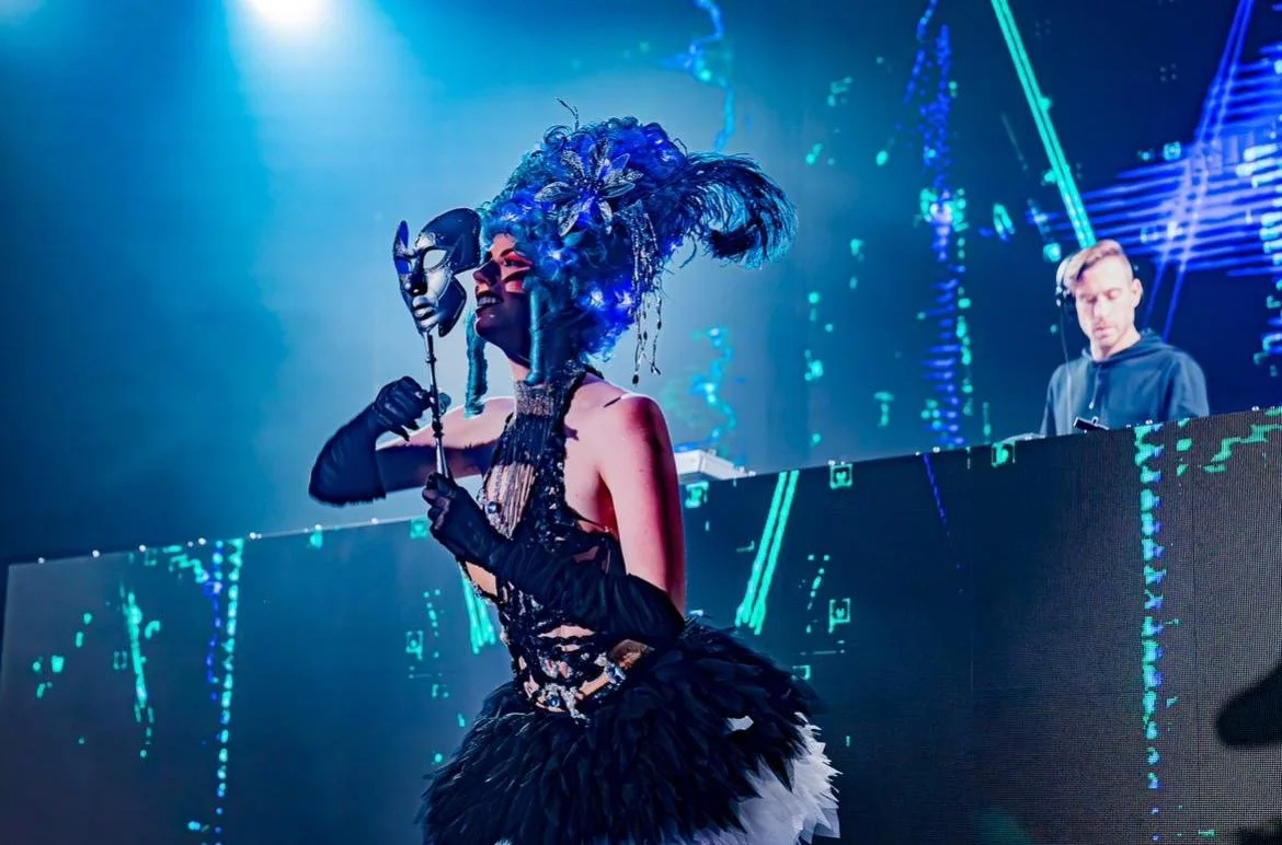 A performer on stage dressed in an elaborate costume with a large feathered headdress, holding a black mask, with a DJ in the background playing music on a digital setup.