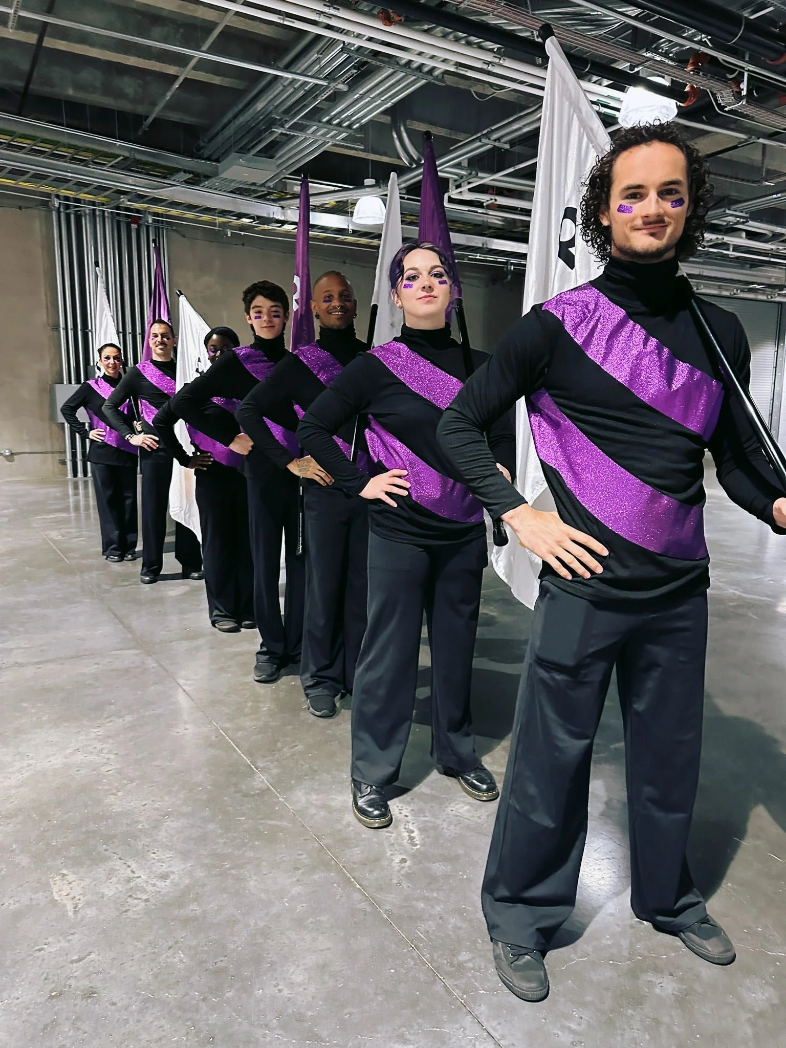 Group of performers in black and purple costumes, standing in a line with flags, backstage or industrial setting.