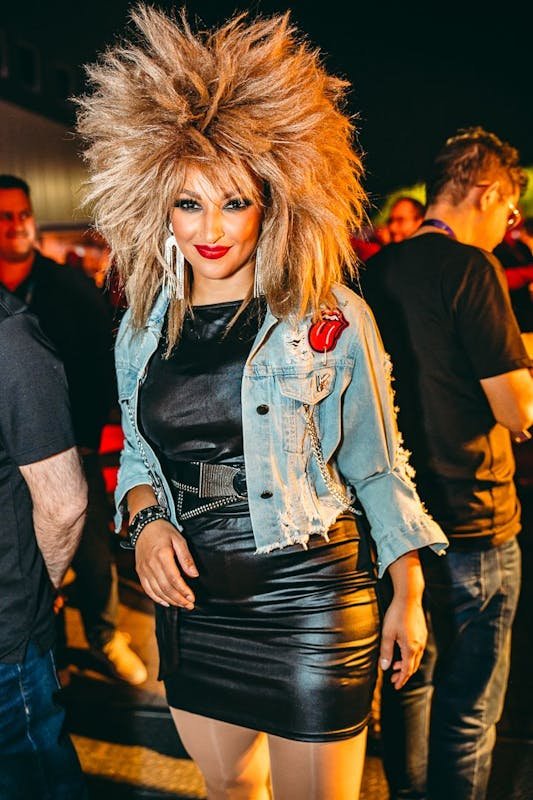 Woman with big, wild blonde hair wearing a denim jacket with Rolling Stones patch, black leather dress, and statement earrings at a lively event.