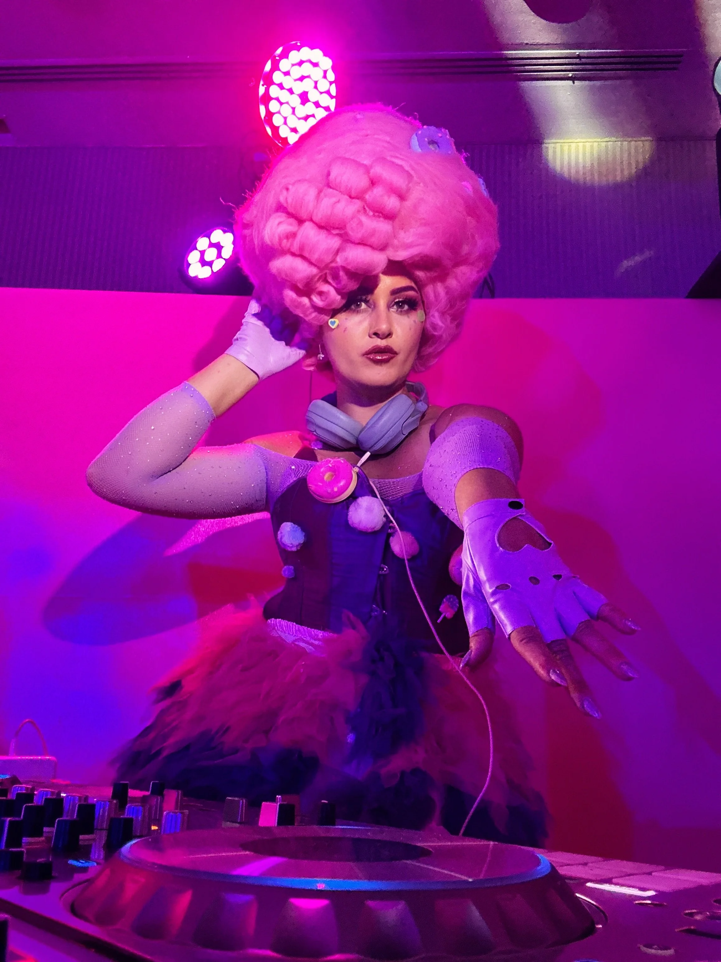 Person dressed in colorful, futuristic costume with pink wig and headphones, posing behind a DJ turntable under purple and pink lights.